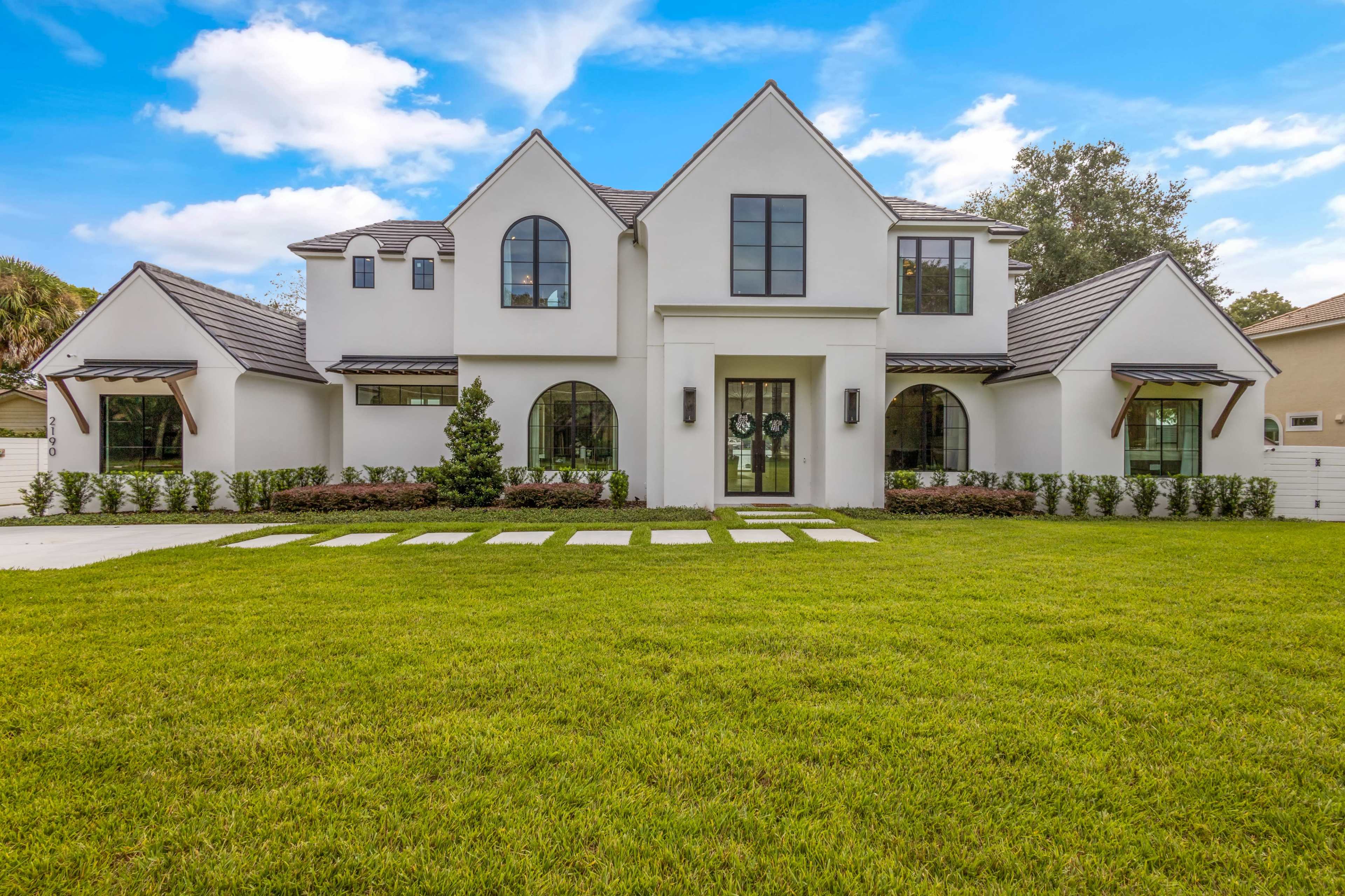 A two-story white house with a black roof features arched windows, a large front porch, and a manicured lawn with stone pathways.