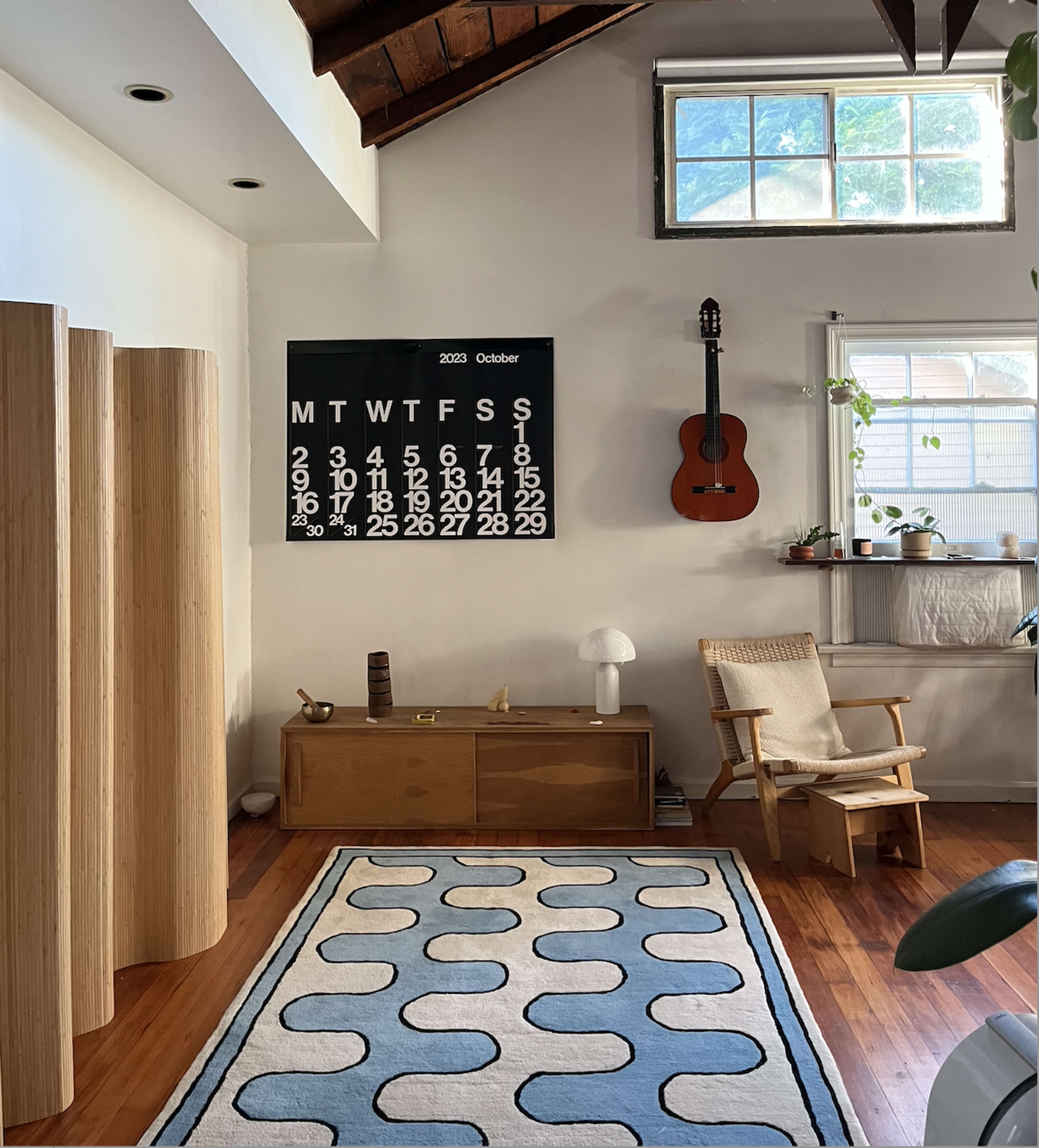 A simple living room features a wooden chair, a calendar on the wall, a guitar hanging, and a modern rug on the floor.