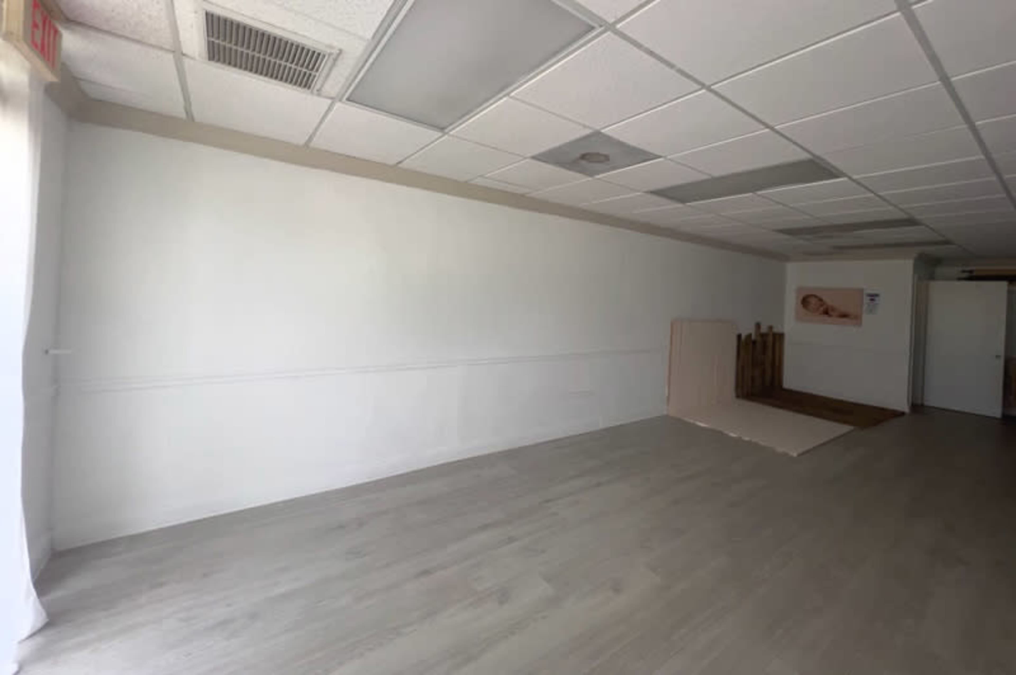 The image shows an empty room with a light-colored floor, a white wall on one side, and a wooden structure partially visible at the back.