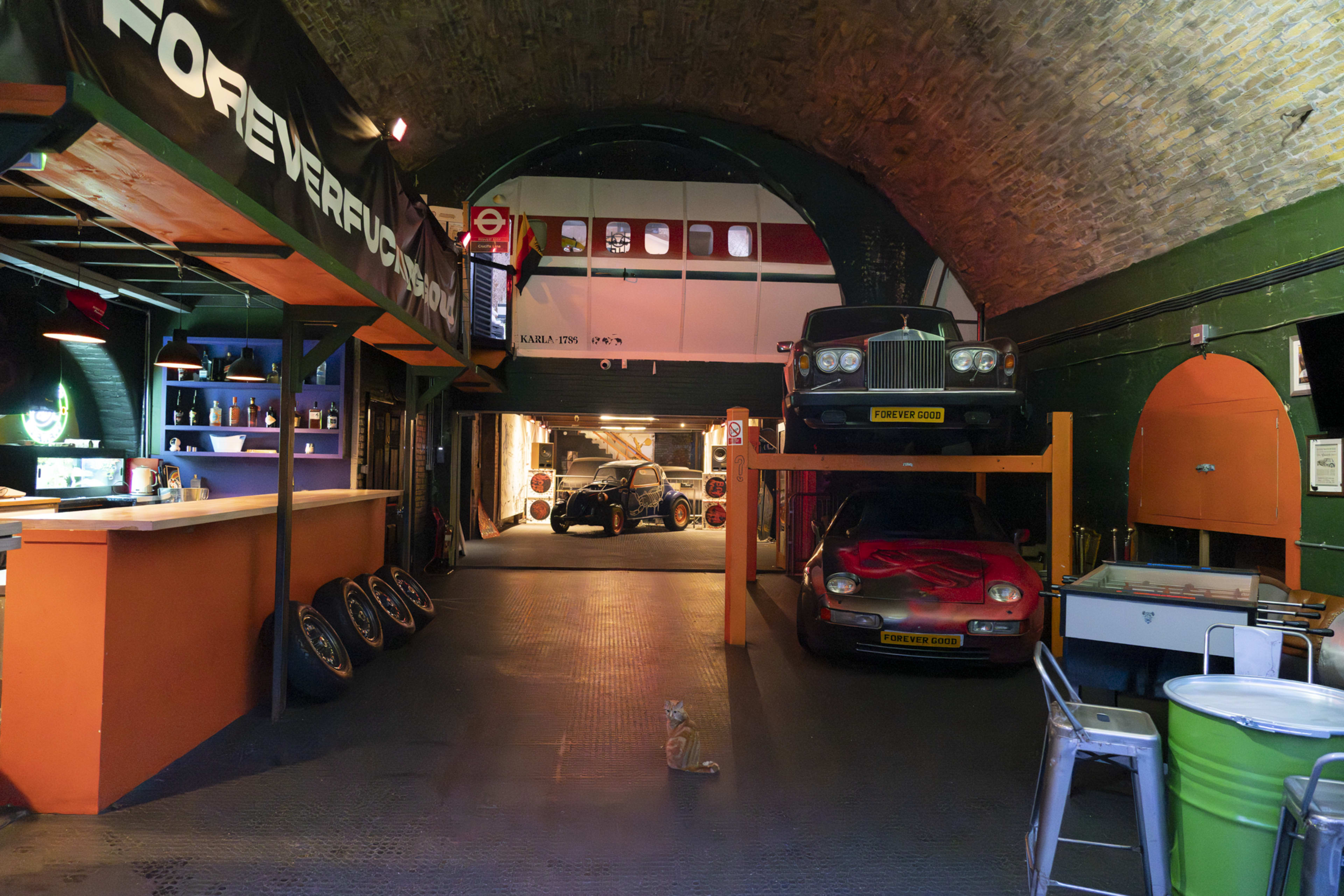 The image depicts an underground garage and bar area featuring classic cars, a vibrant bar with bottles, and a cat sitting on the floor.