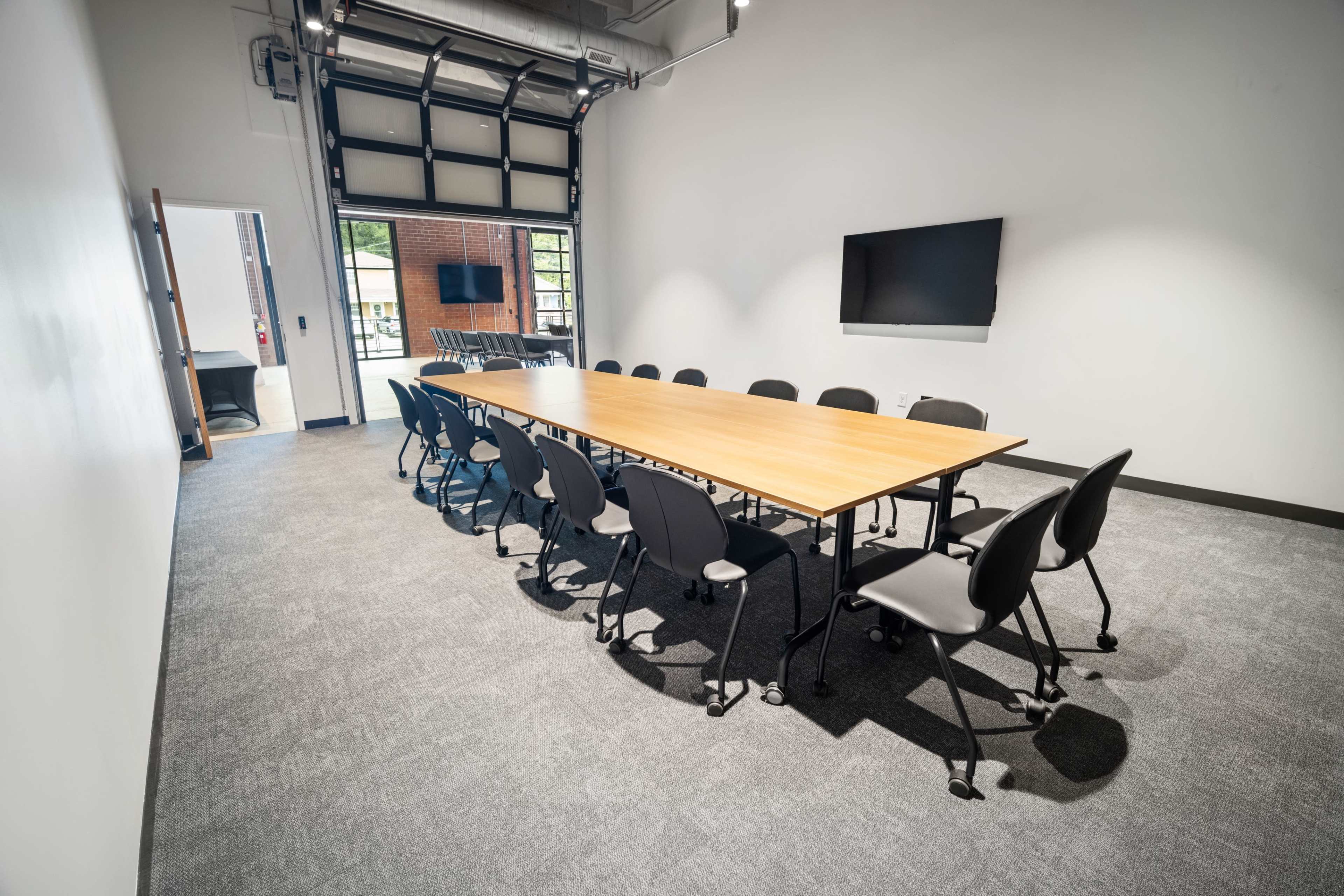 A large conference room features a long wooden table surrounded by black chairs, with a television mounted on the wall and a garage-style door leading to an outside area.