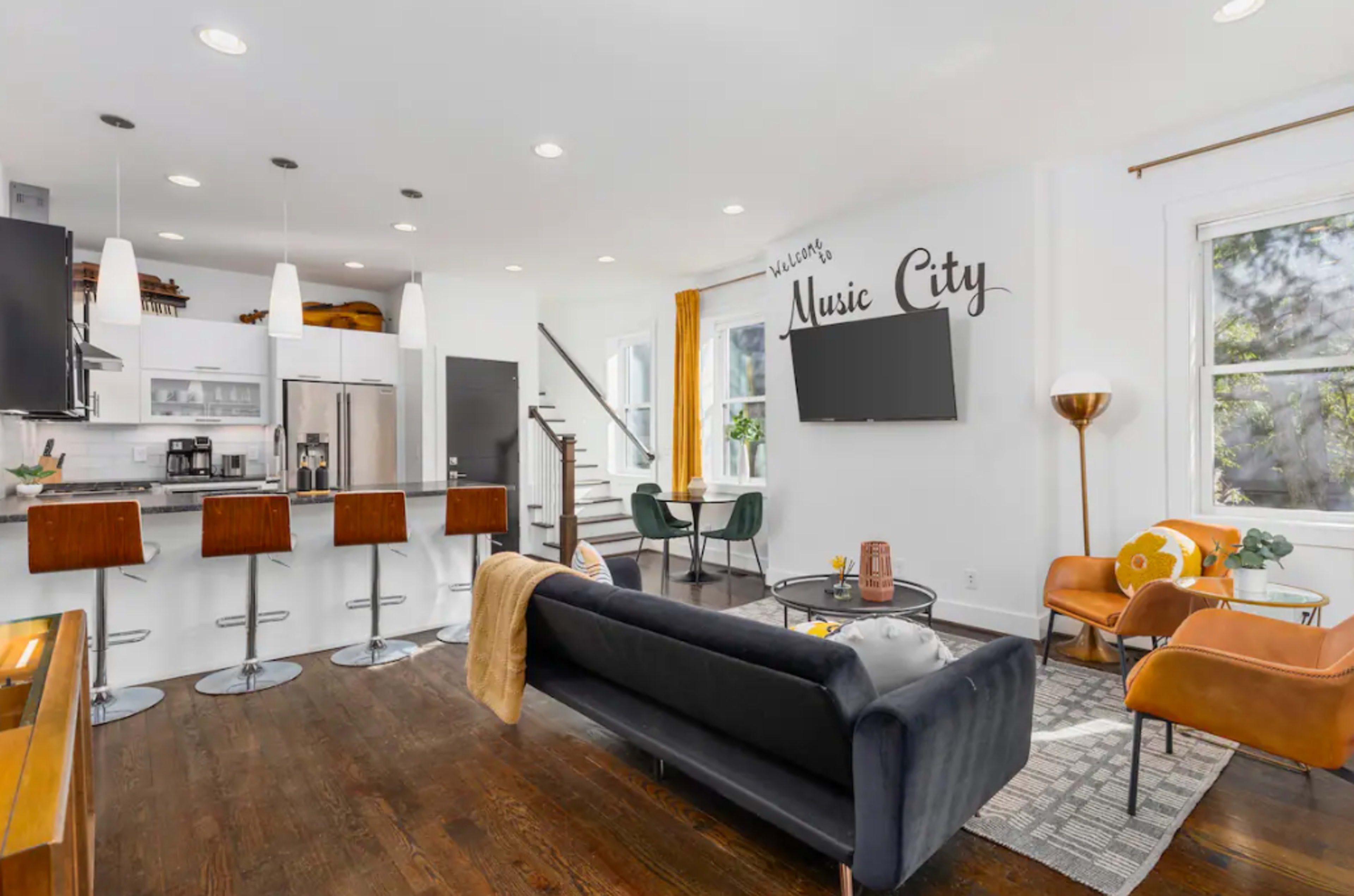 A modern living space with a kitchen area, a seating arrangement featuring a dark sofa and orange chairs, and a staircase in the background.
