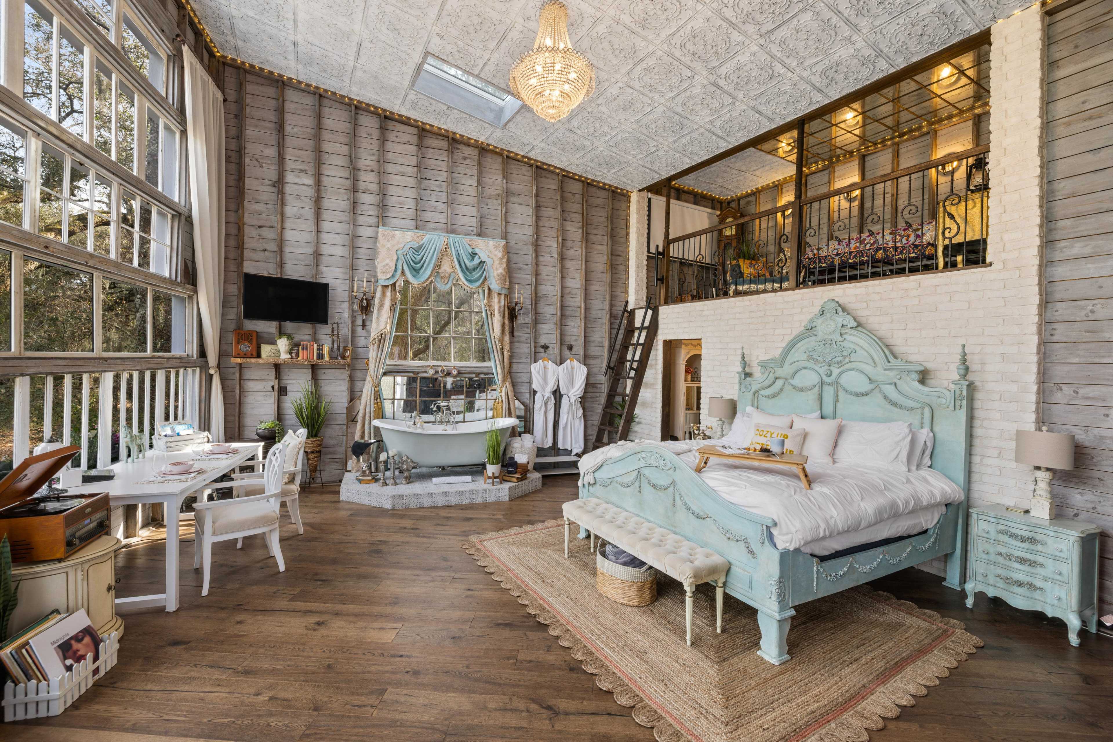 A spacious bedroom with a vintage-style bed and a freestanding bathtub, illuminated by natural light through large windows.