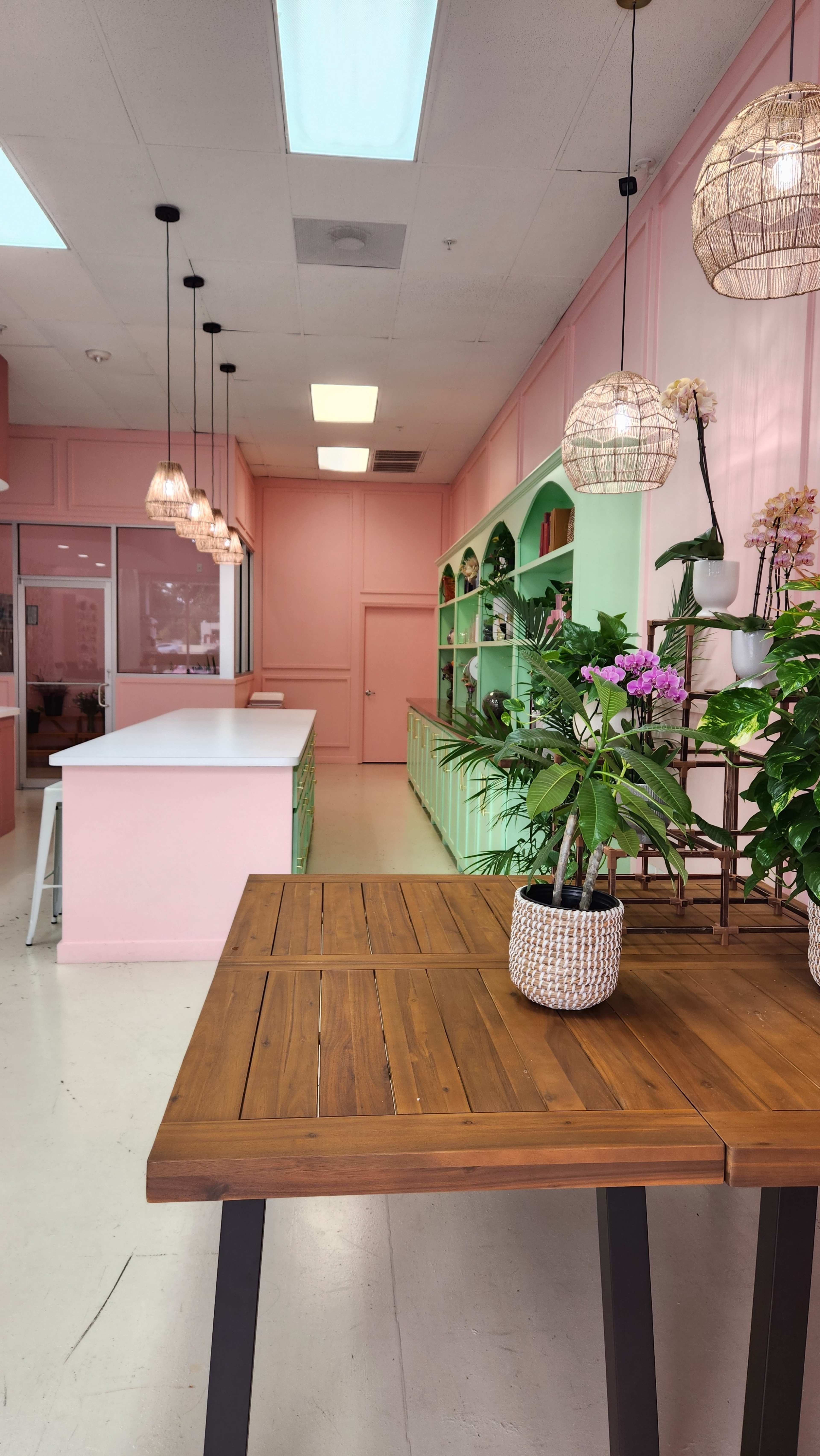 A bright, pastel-colored interior of a shop with a wooden table in the foreground, surrounded by plants and decorative shelves.