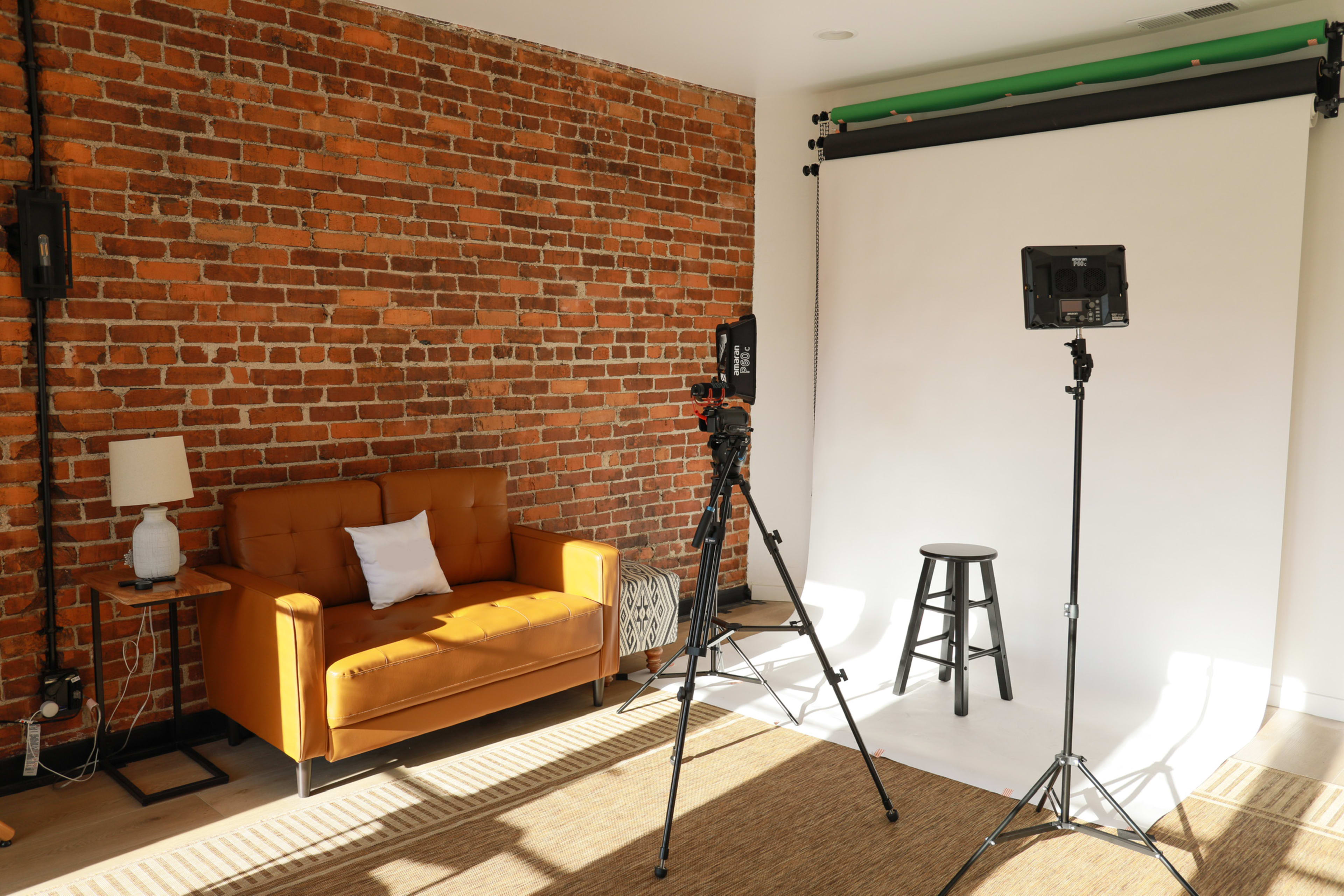 A photography studio with a brick wall backdrop, a brown sofa, a stool, and lighting equipment set up on a white backdrop.