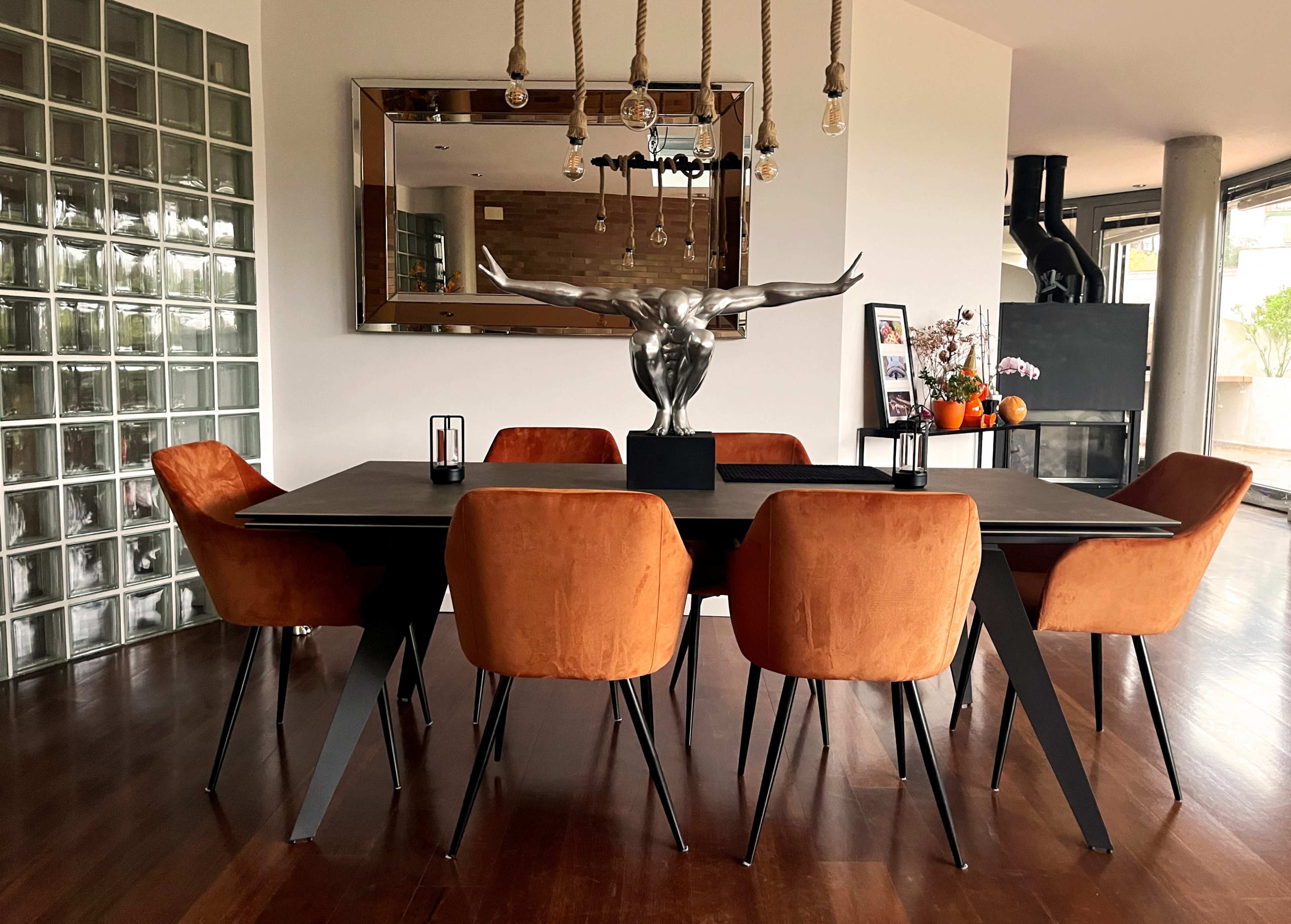 A modern dining room features a large black table surrounded by six orange velvet chairs, with a sculptural centerpiece and a large mirror on the wall.