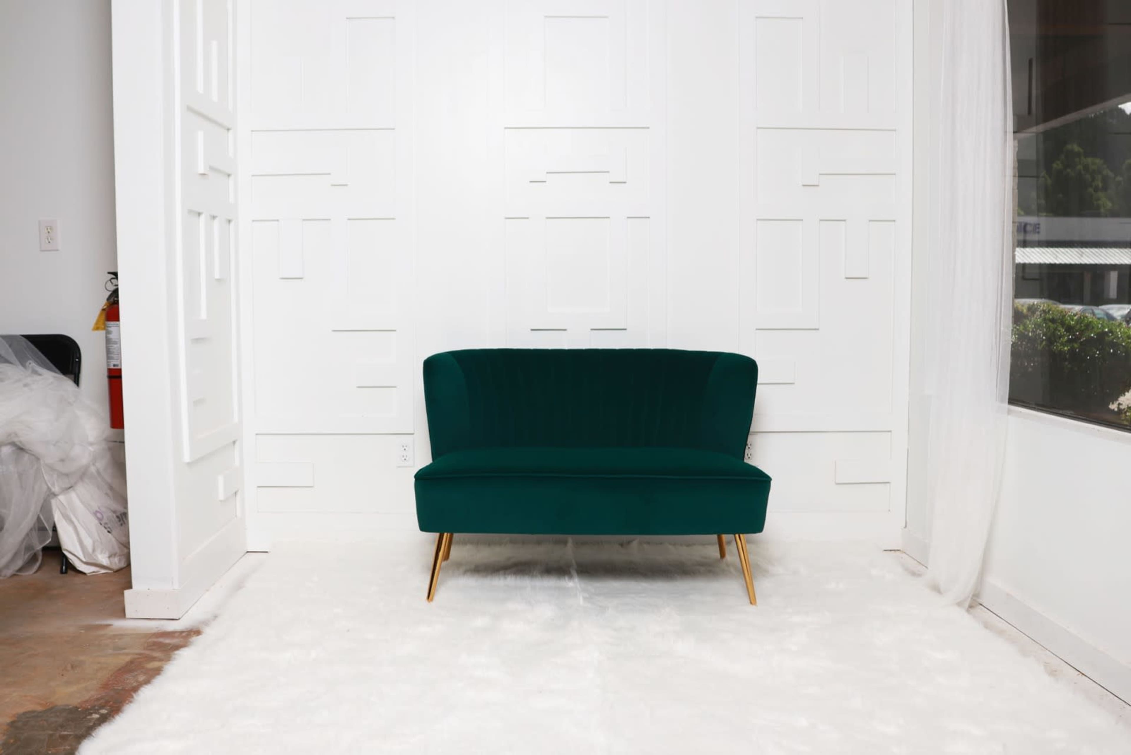 A green velvet loveseat with gold legs is placed on a white rug in front of a white wall with a geometric design.