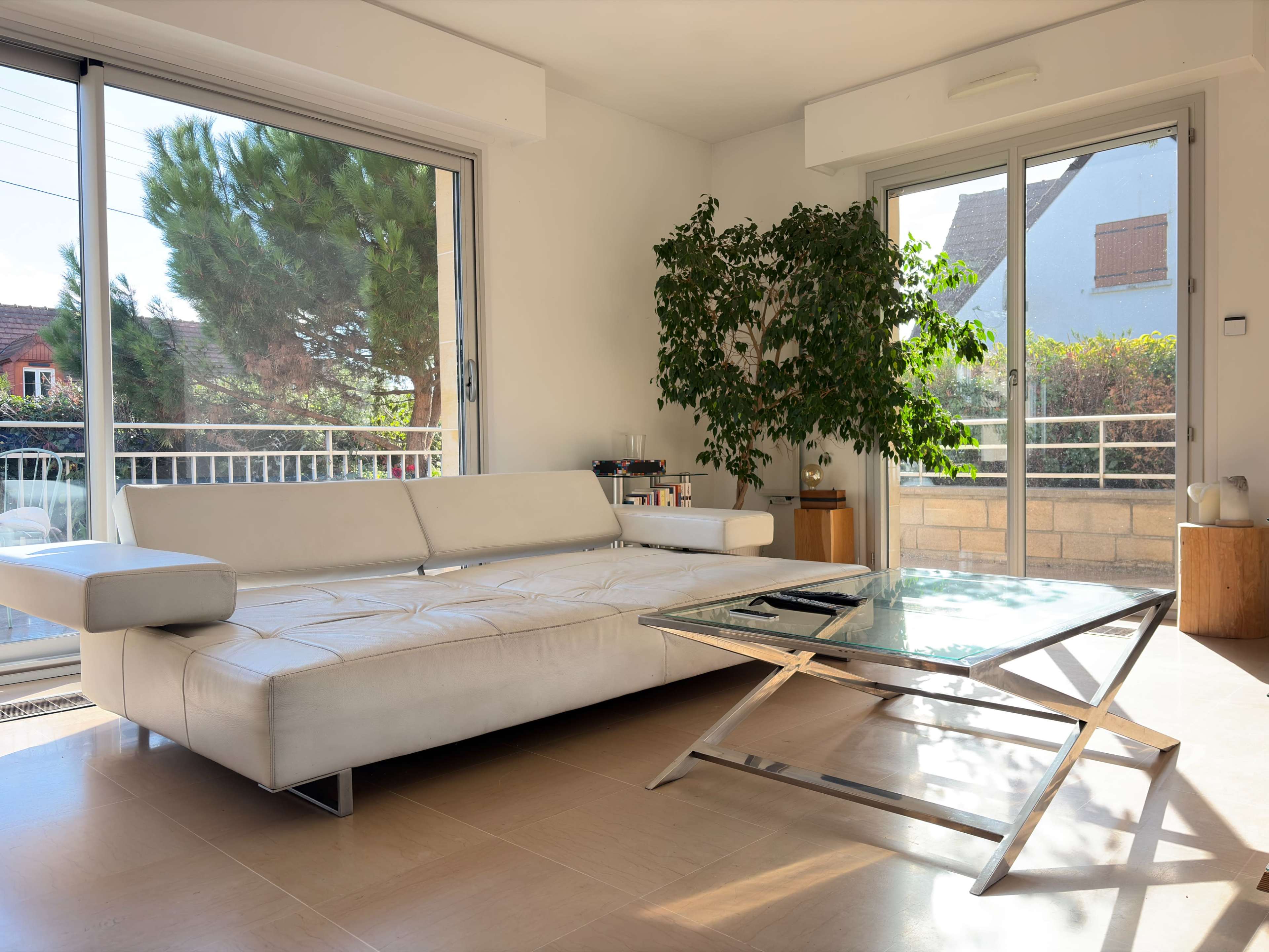 The image shows a modern living room featuring a white sectional sofa, a glass coffee table, and large windows that let in natural light.
