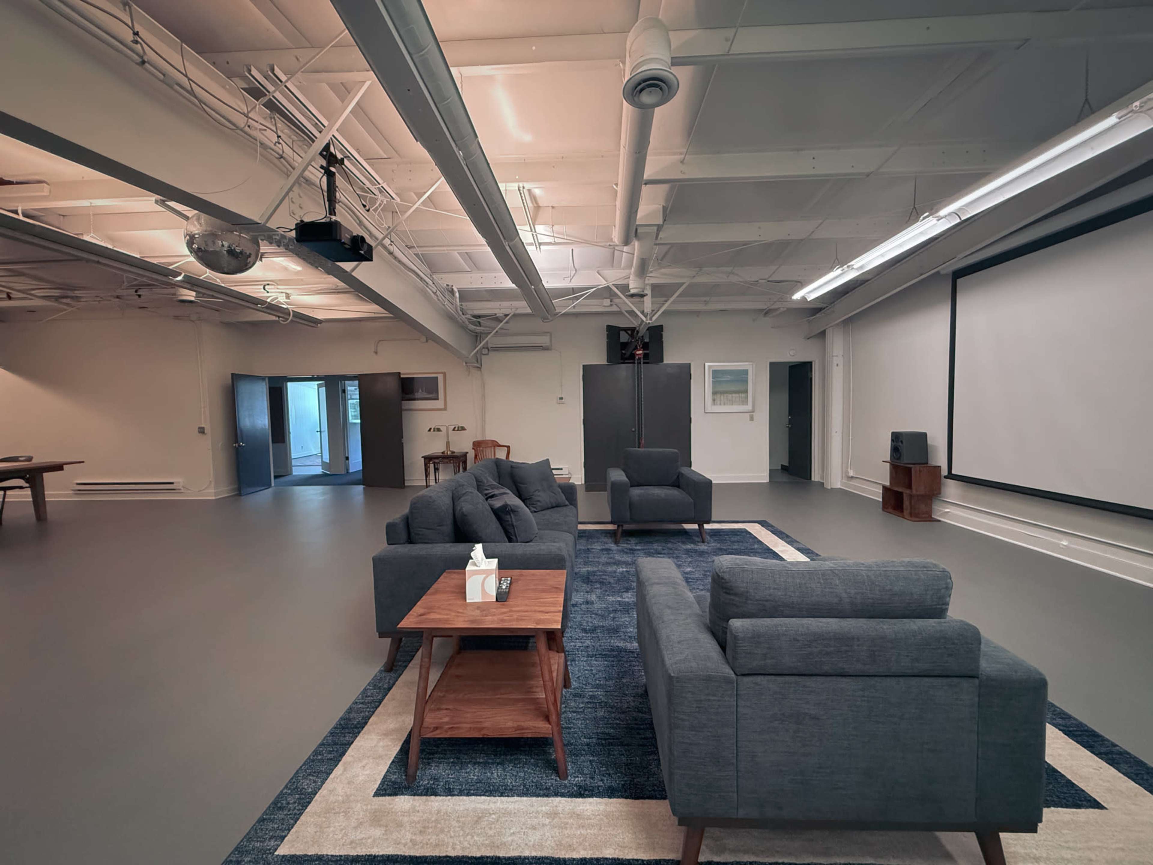 The image shows a spacious room with a seating area consisting of gray sofas and a wooden coffee table, along with a projector screen and large windows.