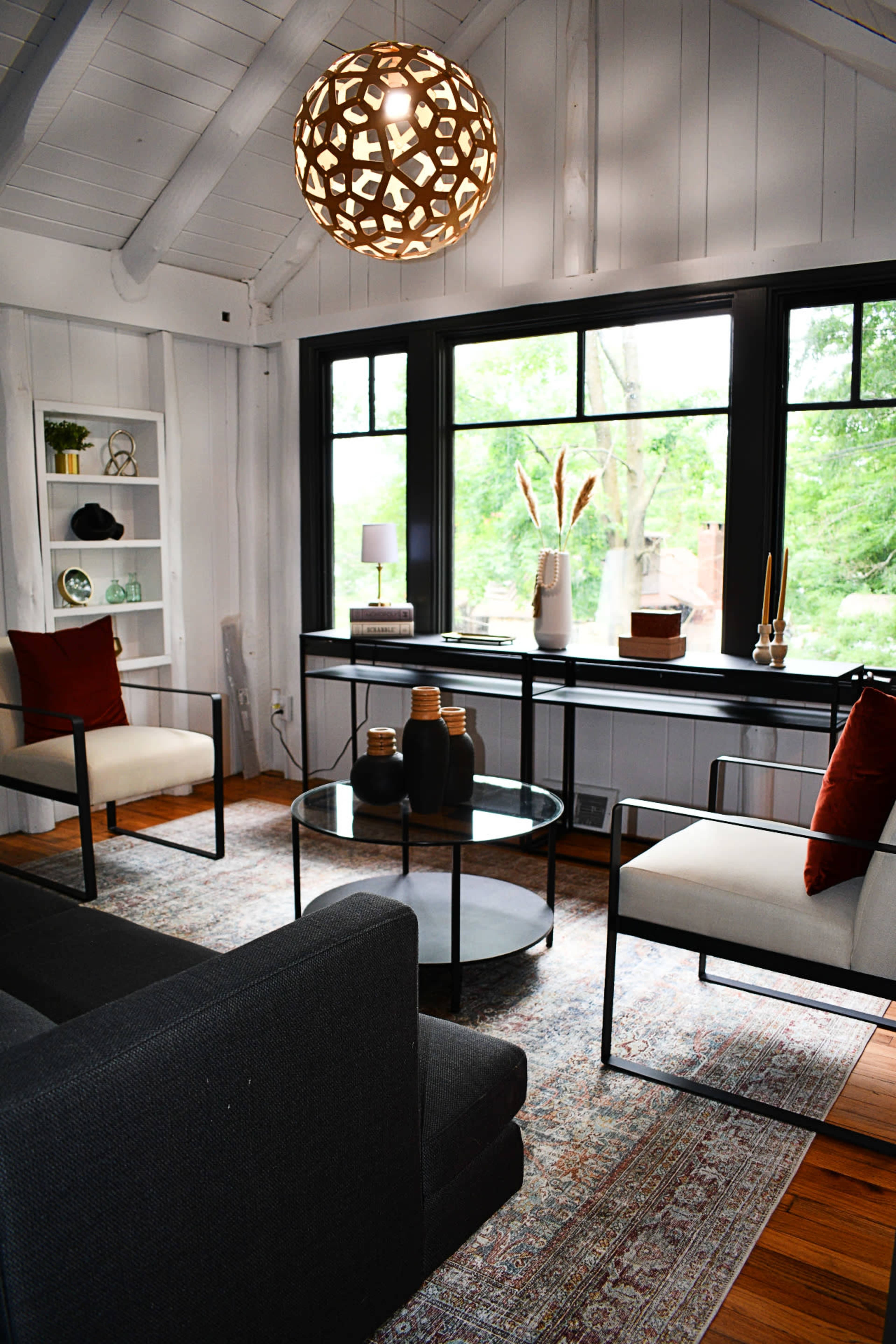 A modern living room featuring two armchairs, a round glass coffee table, and a decorative light fixture, with large windows allowing natural light to illuminate the space.