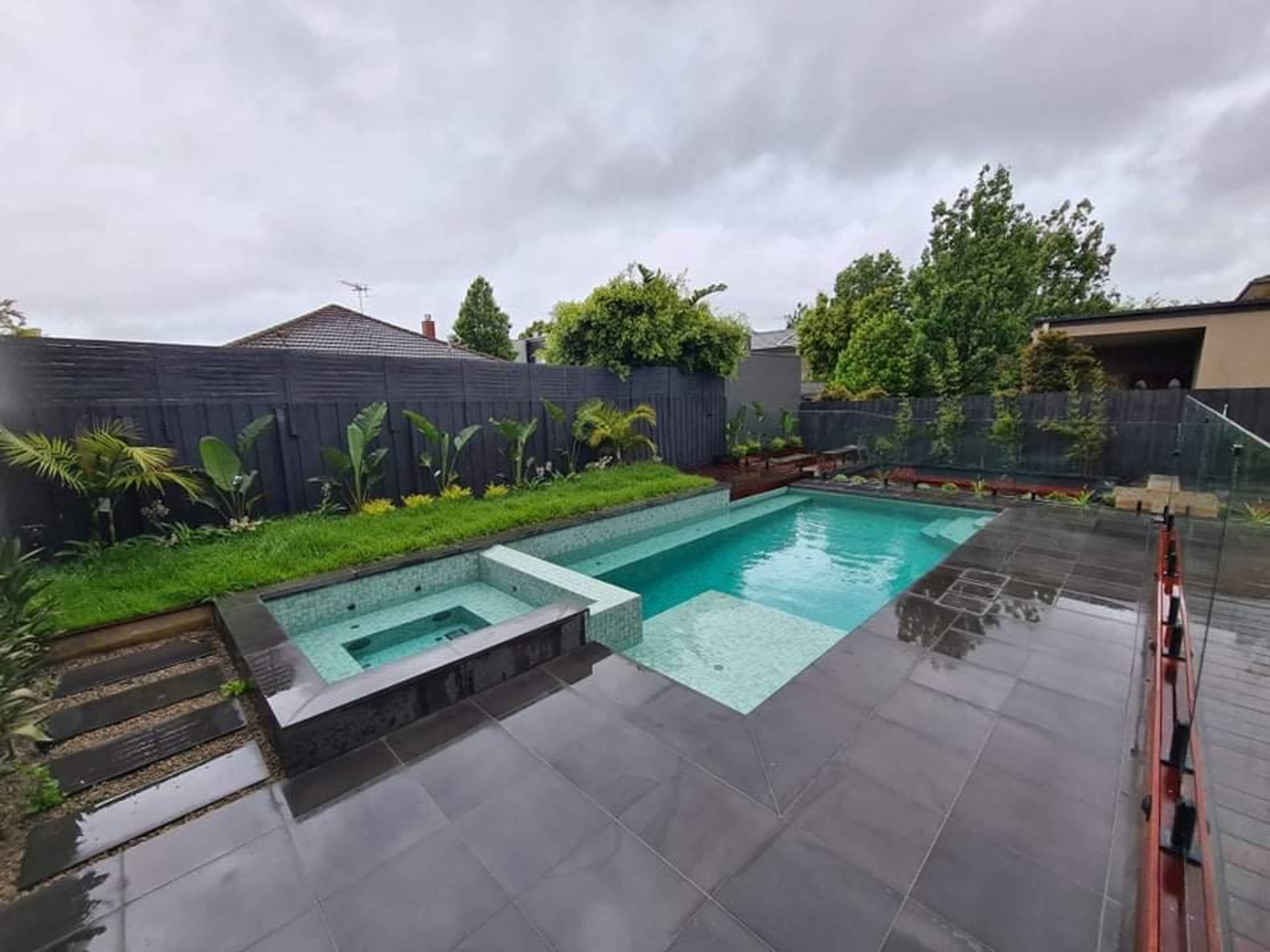 A modern backyard featuring a rectangular swimming pool, a spa area, and landscaped greenery surrounded by a wooden fence.