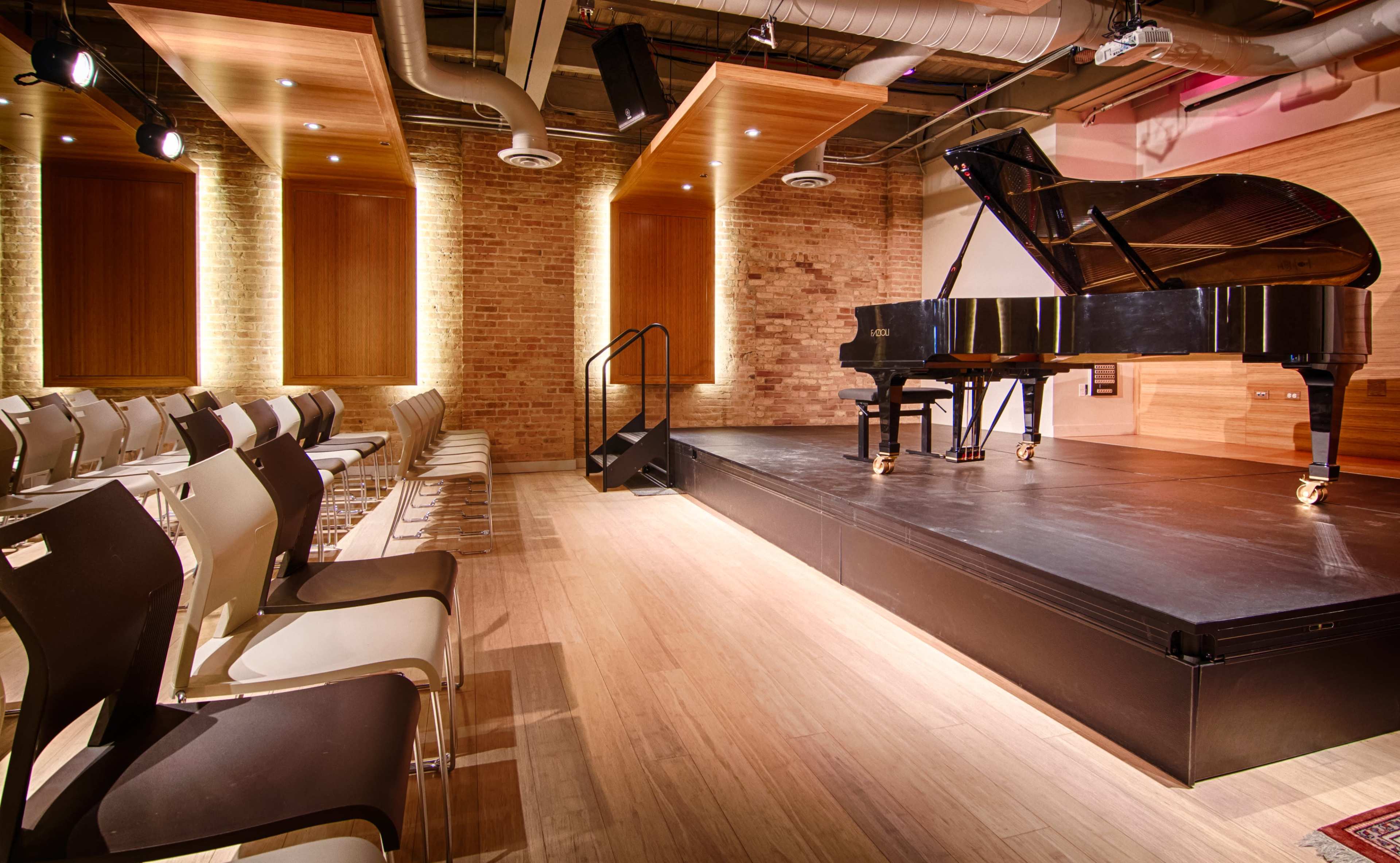 A performance space featuring a grand piano on a raised platform with empty chairs arranged in front of it.