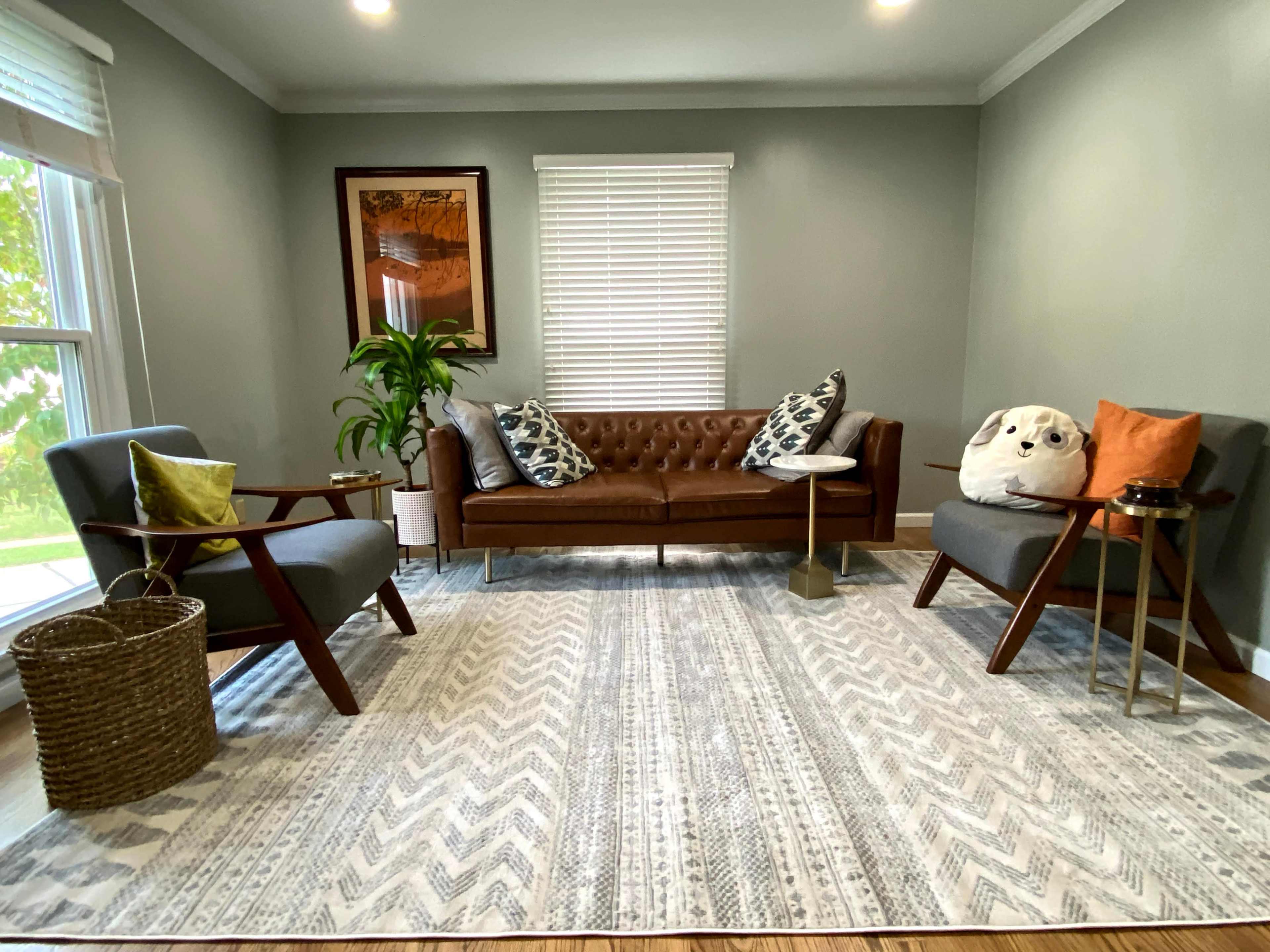 A cozy living room with a brown leather sofa, two armchairs, a patterned area rug, and a window letting in natural light.