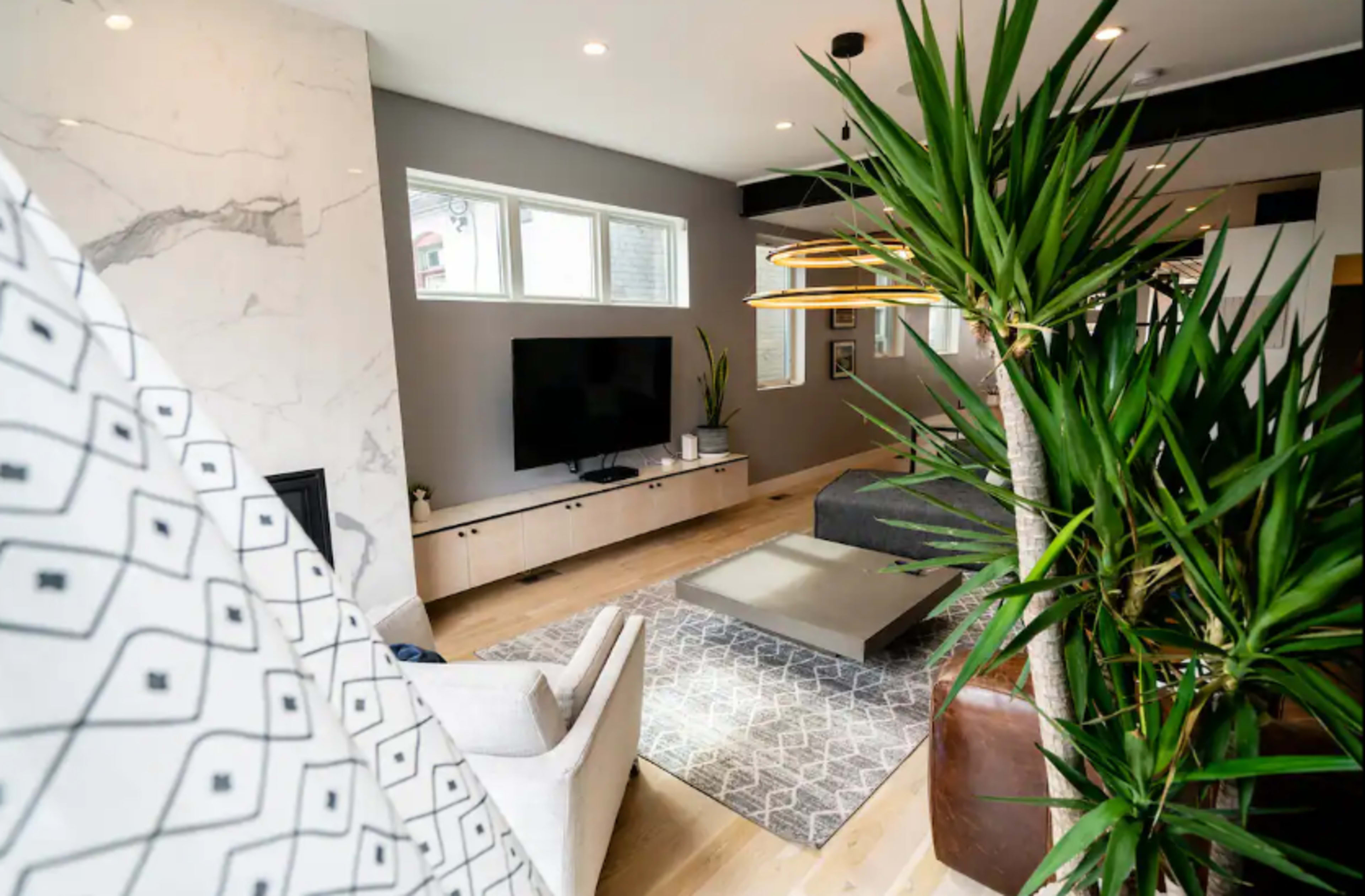 The image shows a modern living room featuring a television against a light wooden unit, a gray sofa, and several plants, with large windows allowing natural light to enter the space.