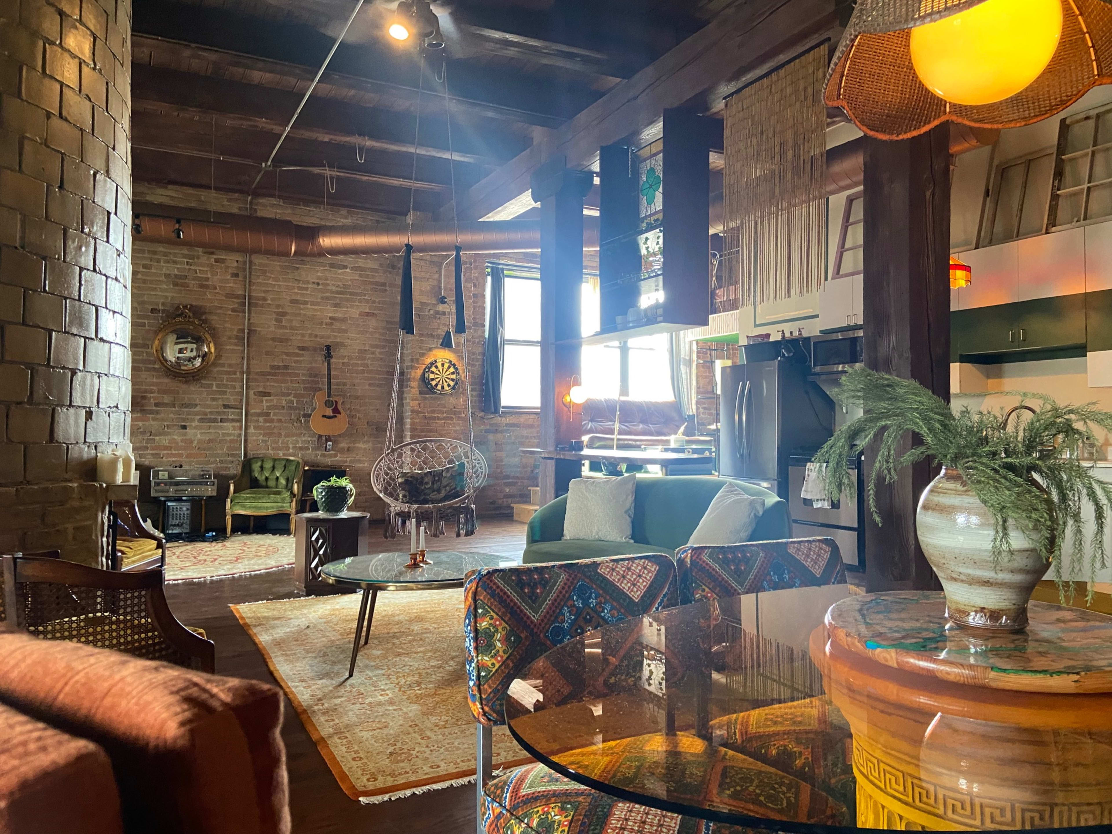 A cozy living space featuring a mix of vintage furniture, decorative items, and large windows, with a guitar and a dartboard on the walls.