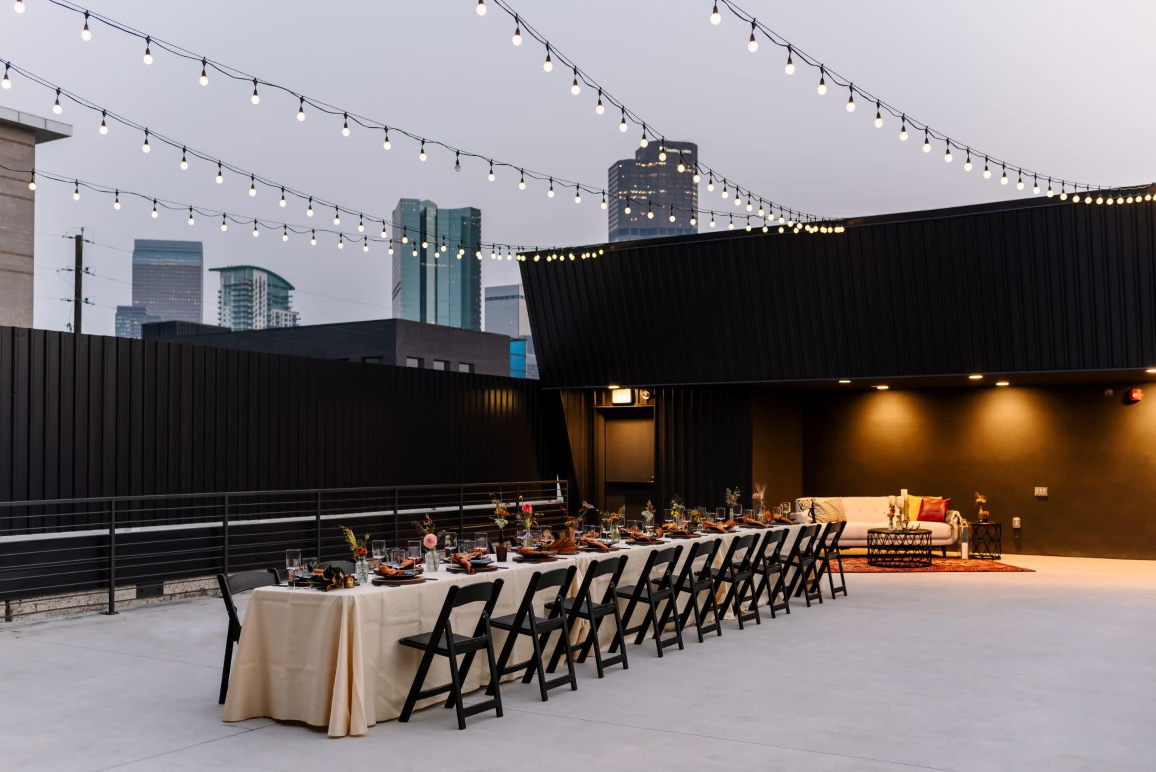A long table set for dinner is arranged under string lights on a rooftop with city skyline views.