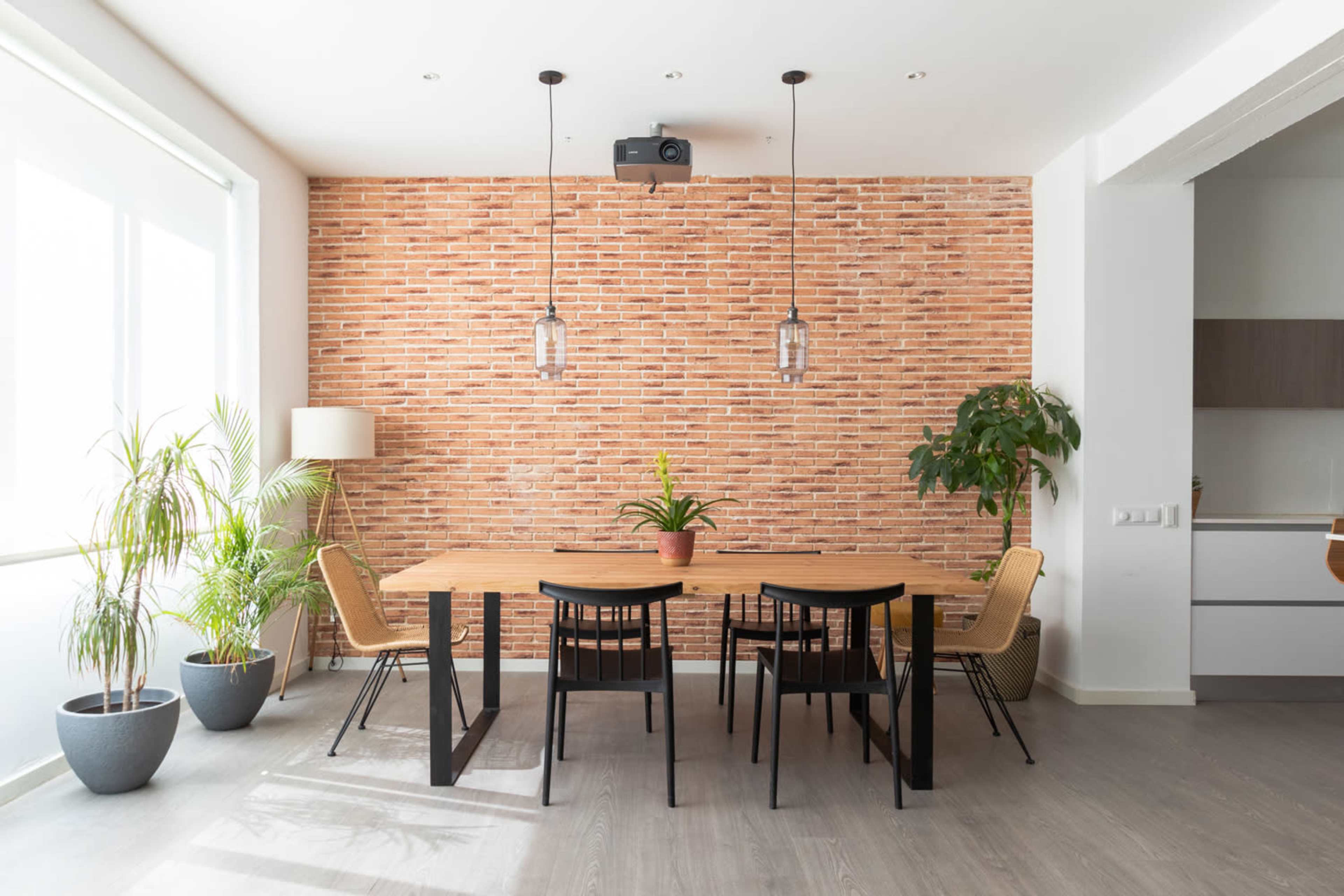 A dining area features a wooden table surrounded by black chairs, set against a brick wall with plants and a projector overhead.