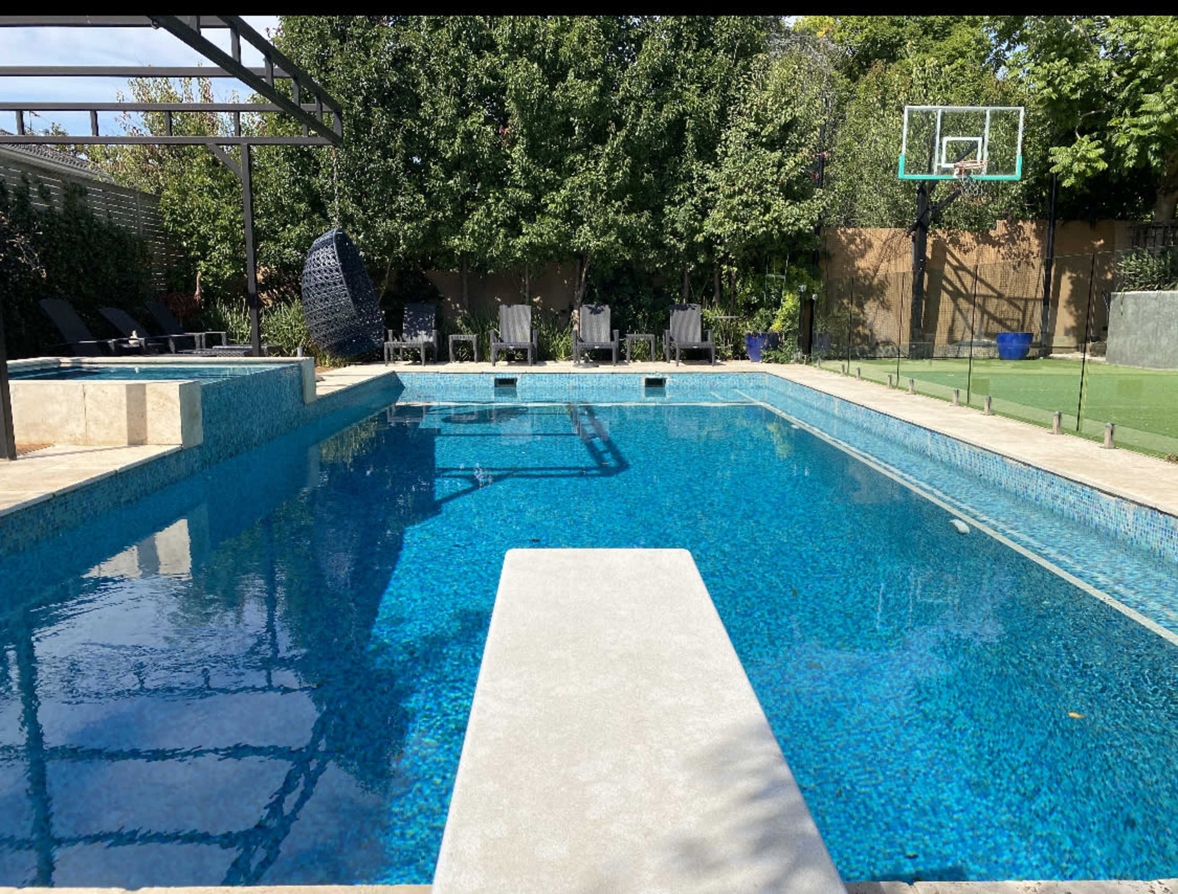 A rectangular swimming pool with a diving board, surrounded by lounge chairs and a basketball hoop in a landscaped backyard.