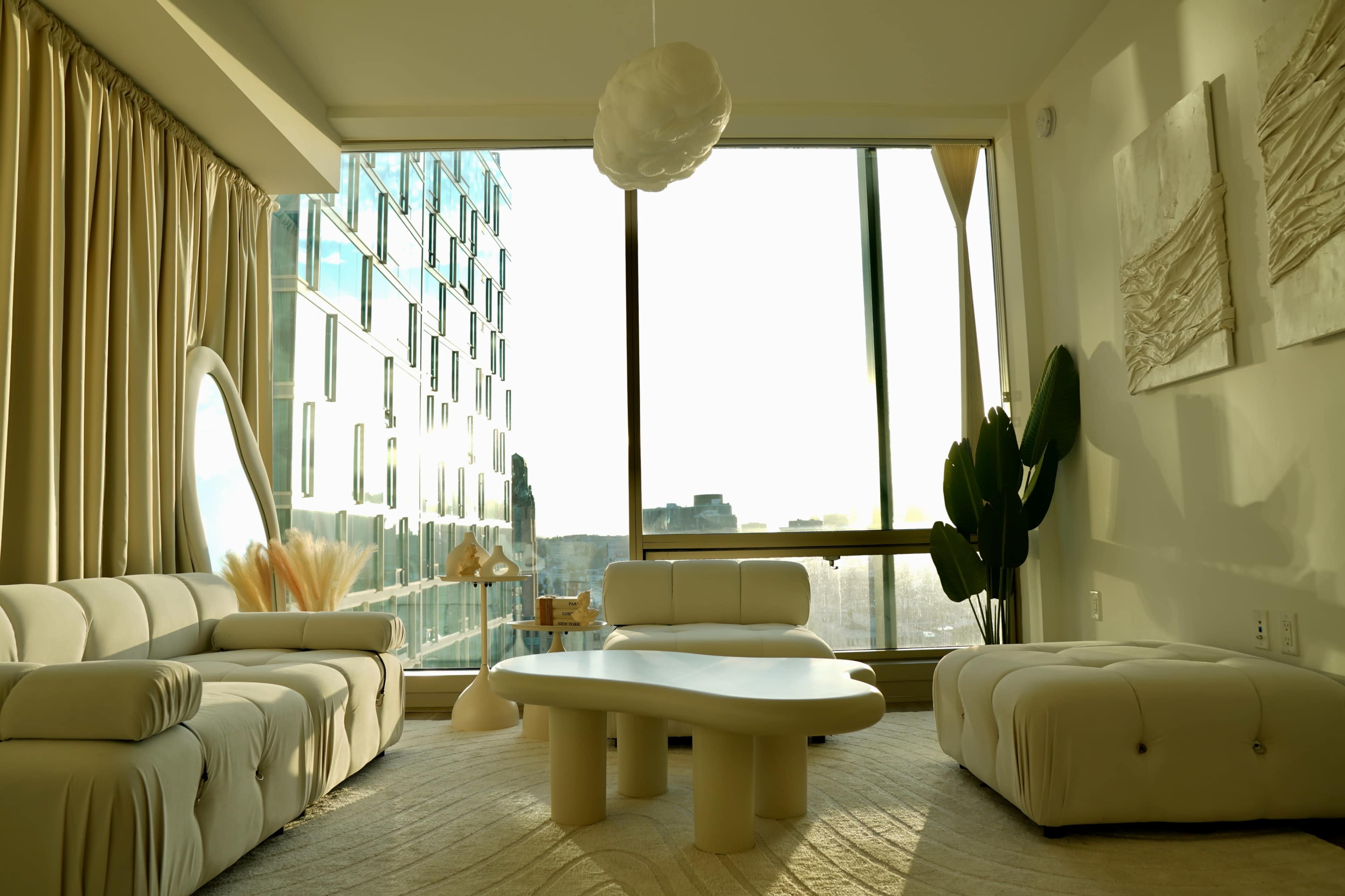 A modern living room features a white sectional sofa, a rounded coffee table, and large windows offering a view of the city skyline.