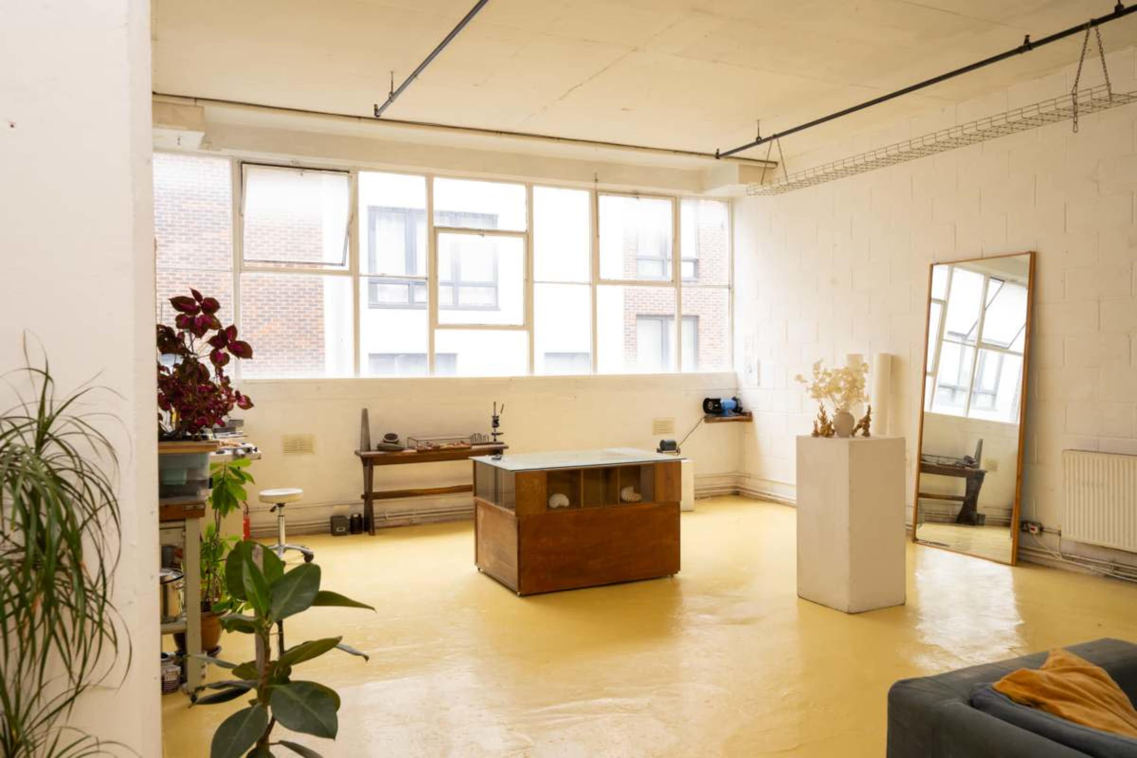 The image shows a bright, spacious studio with large windows, a wooden table in the center, a mirror against the wall, a sofa, and various plants.
