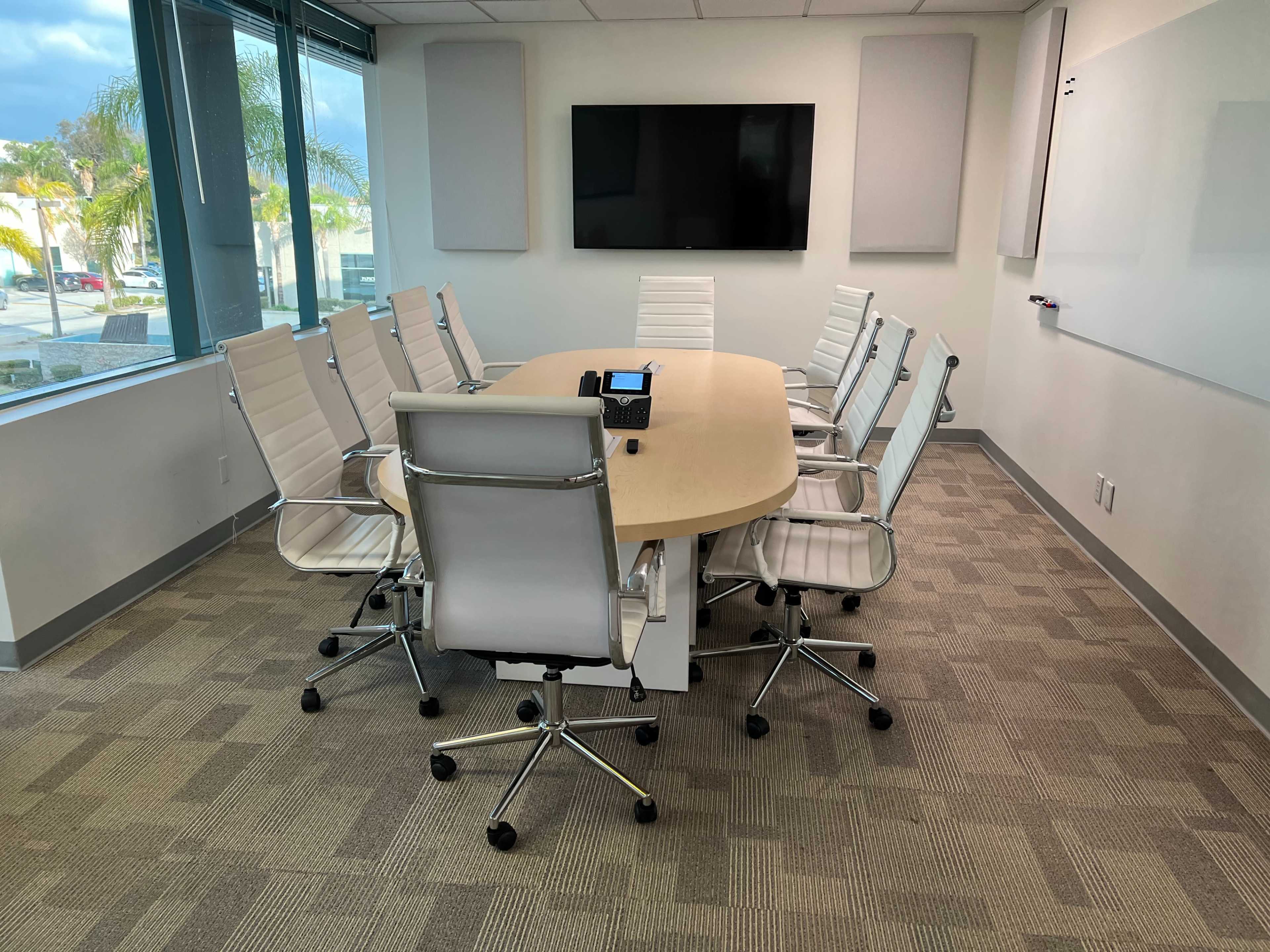 A modern conference room features an oval table surrounded by eight white ergonomic chairs, with large windows and a mounted screen on the wall.