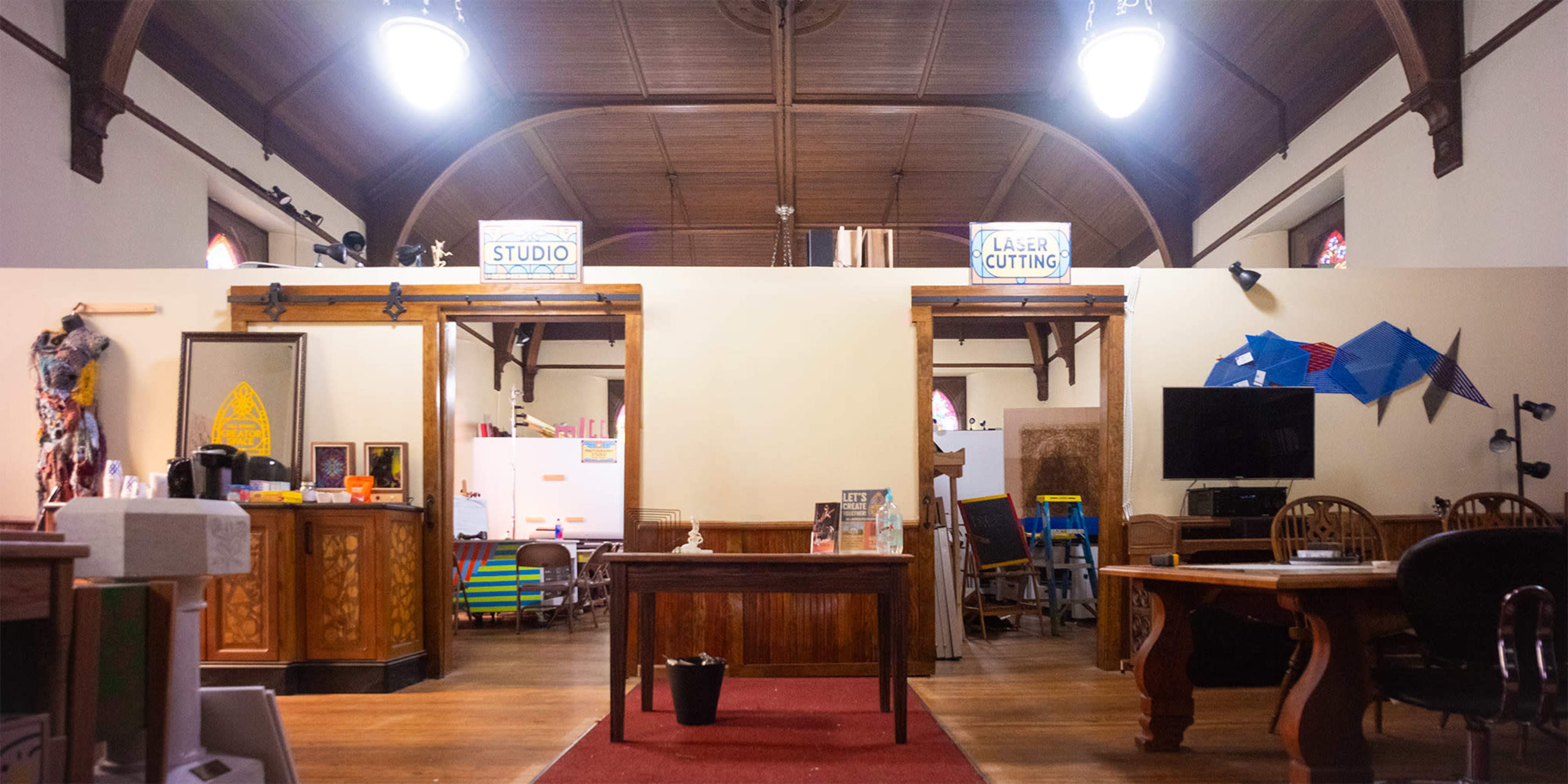 The image shows a spacious workshop with wooden beams, various workstations, and signs for "Studio" and "Laser Cutting" hanging above two entryways.