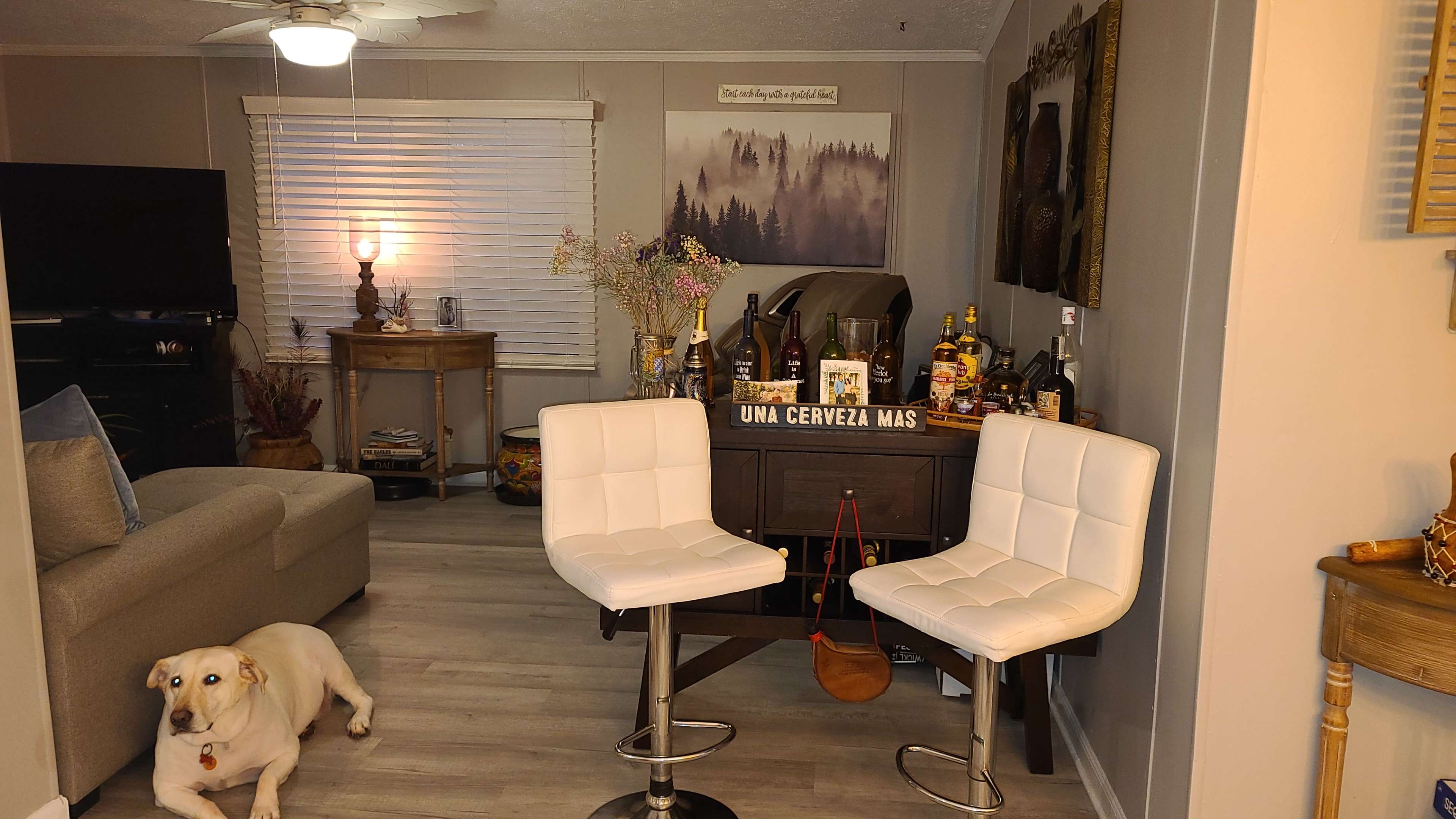 The image shows a cozy living space featuring a beige couch, two white bar stools, a bar cart stocked with various bottles, and a yellow dog lying on the floor.