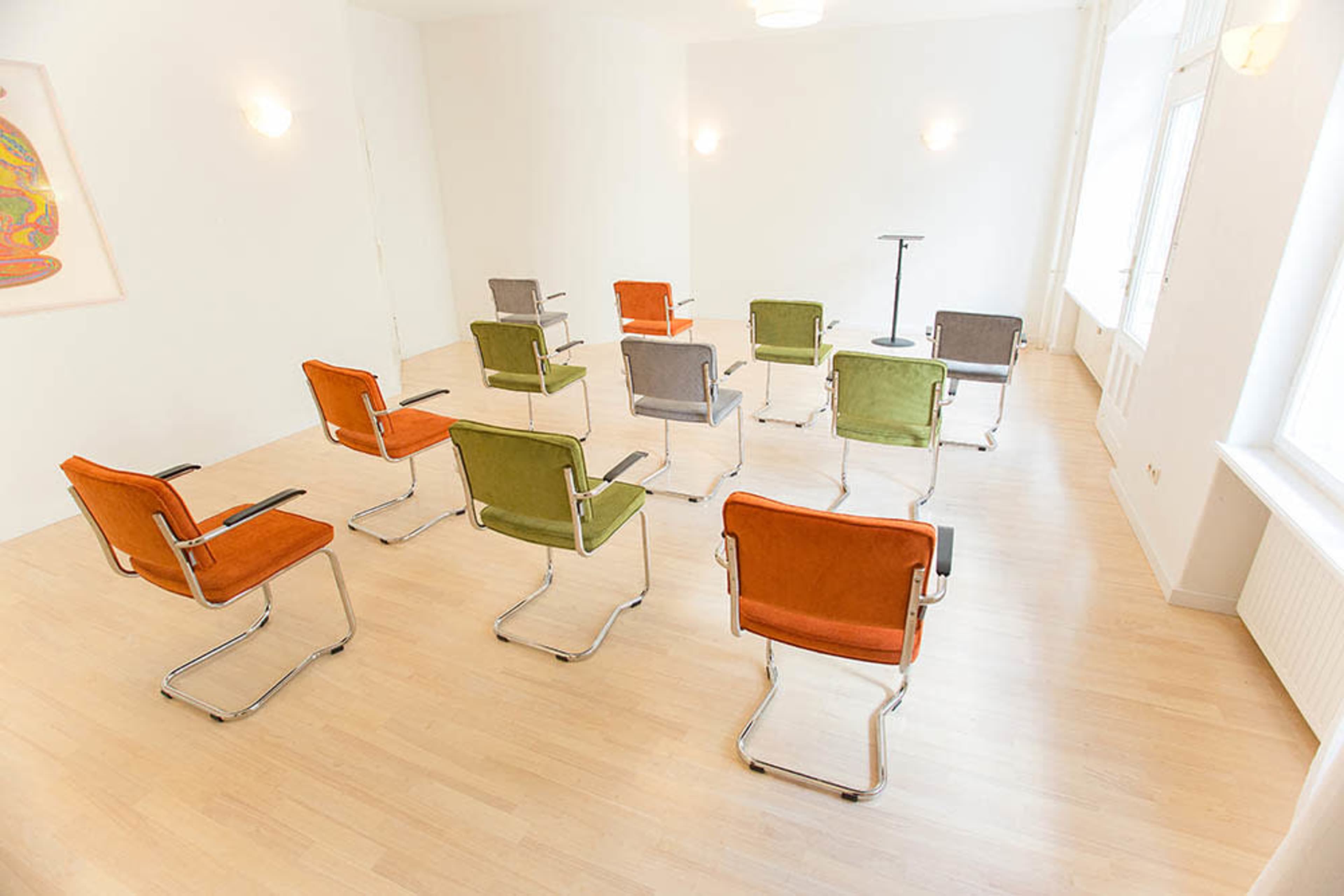 A sparse room features a circular arrangement of ten colorful chairs with a small podium at the center.
