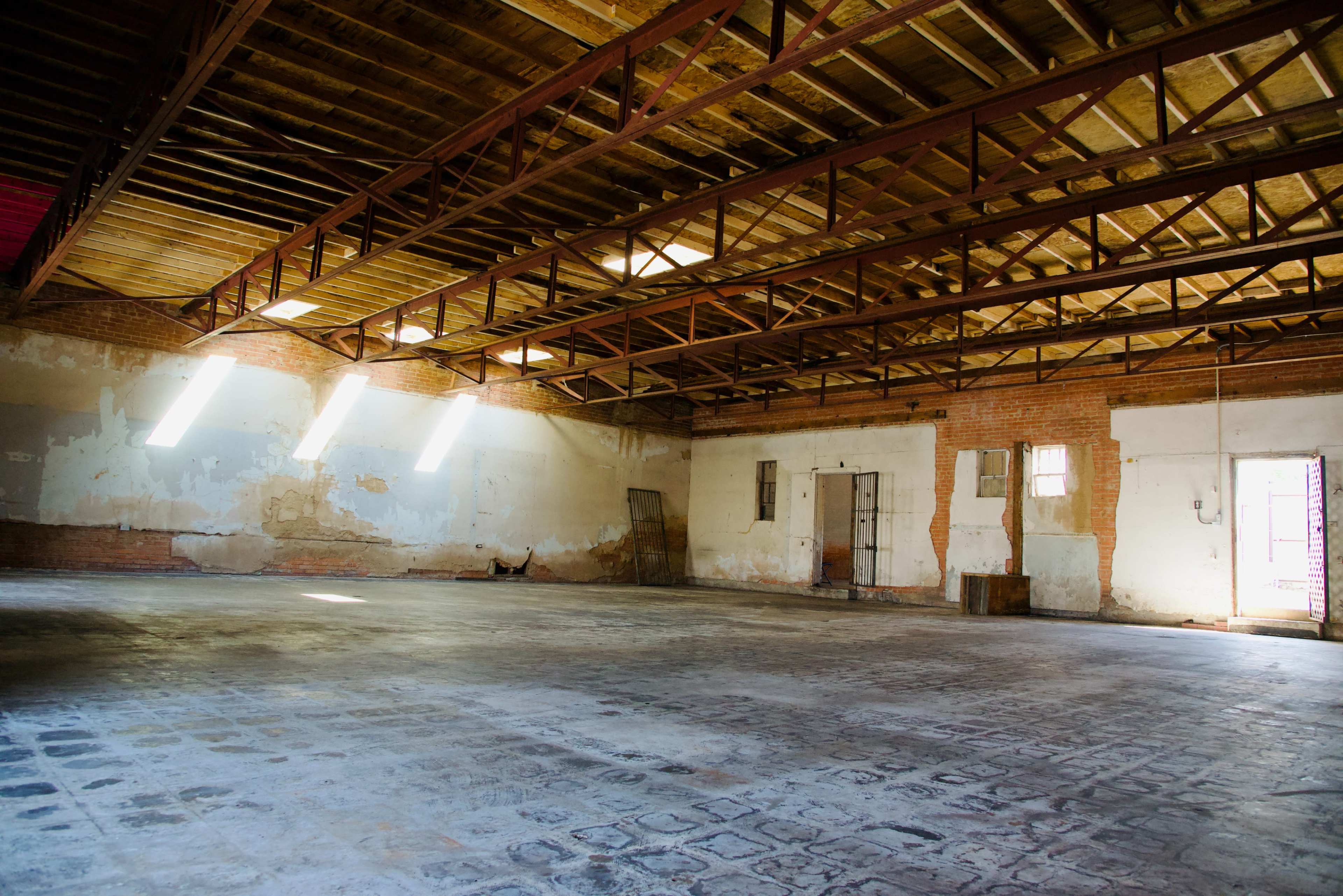 A spacious, empty industrial room with exposed wooden beams and sunlight filtering through large windows onto a bare floor.