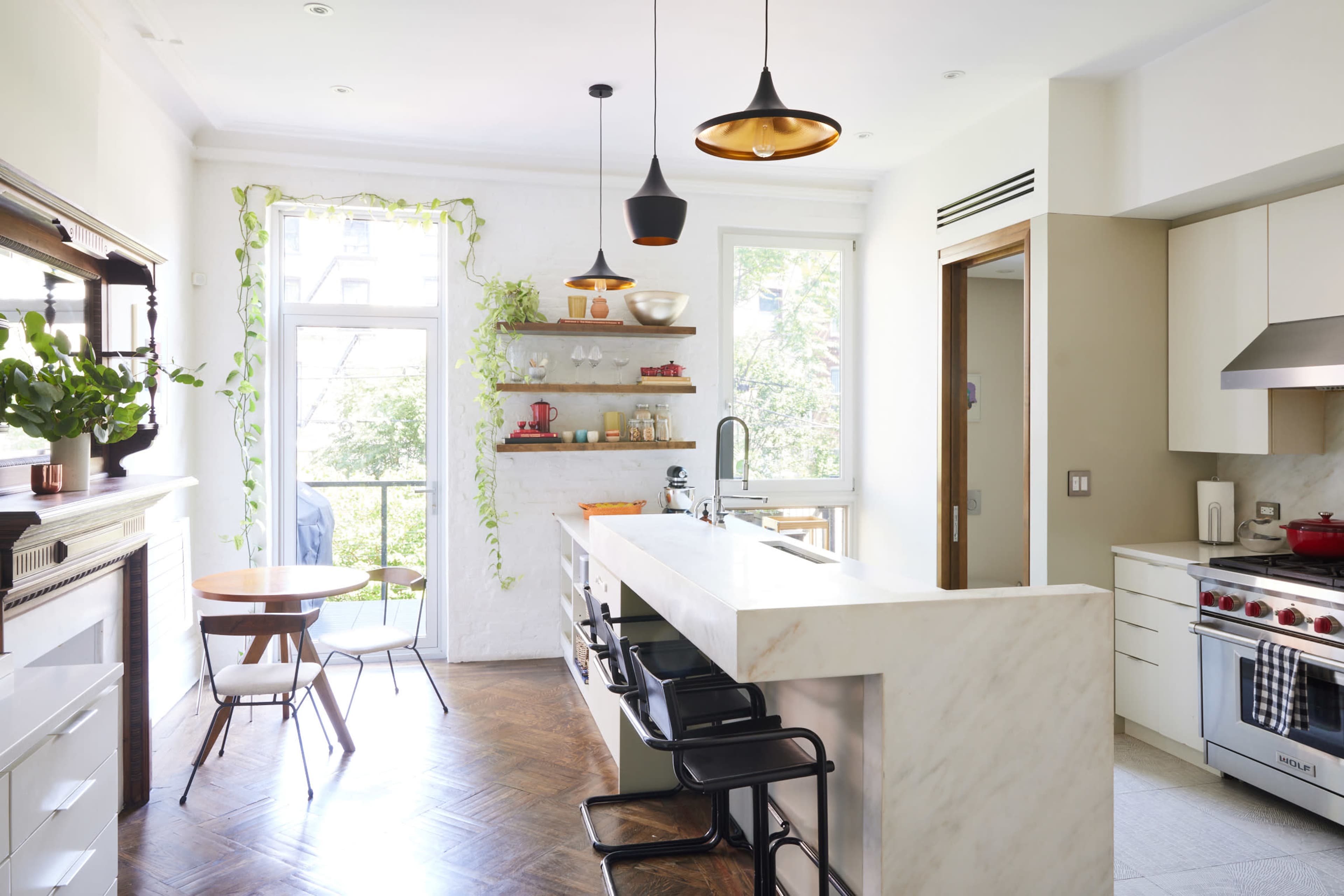 A modern kitchen with a marble island, wooden shelves displaying dishware, and a table by a window.