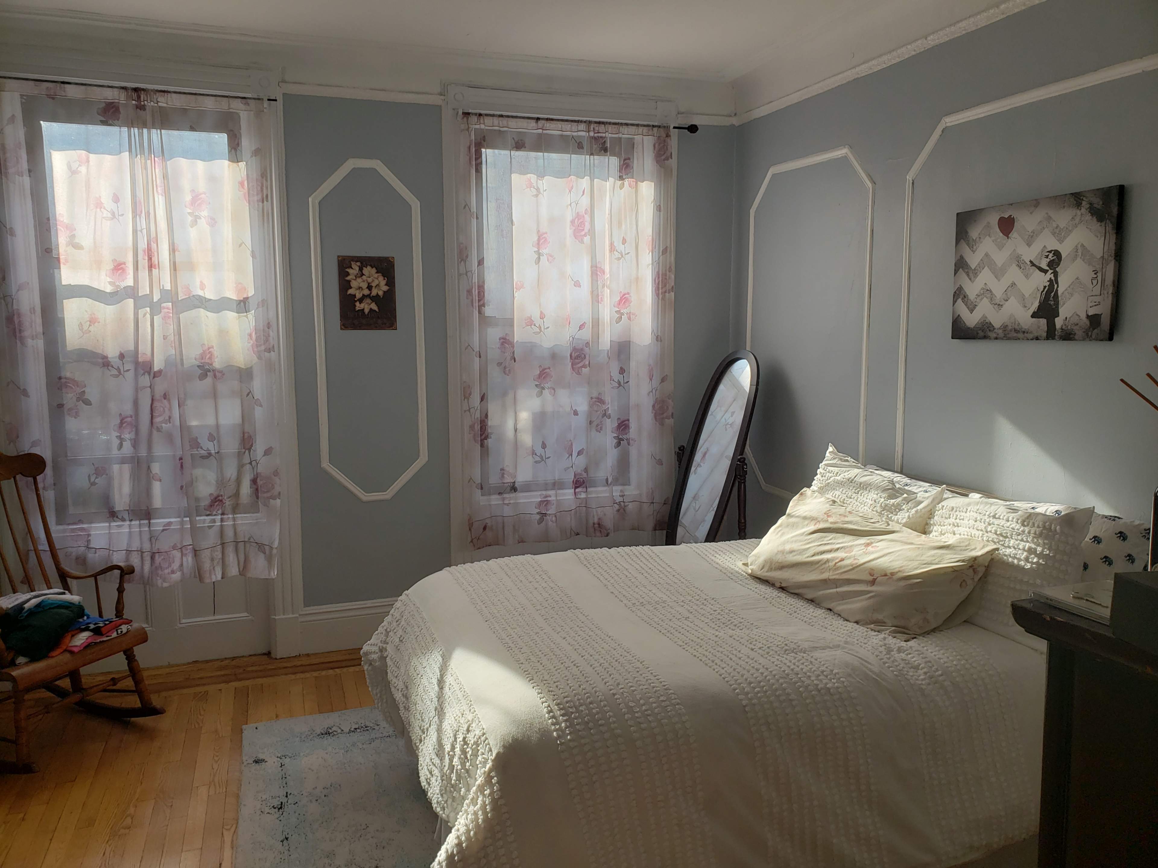A softly lit bedroom with a bed, a rocking chair, and two window treatments featuring floral patterns.