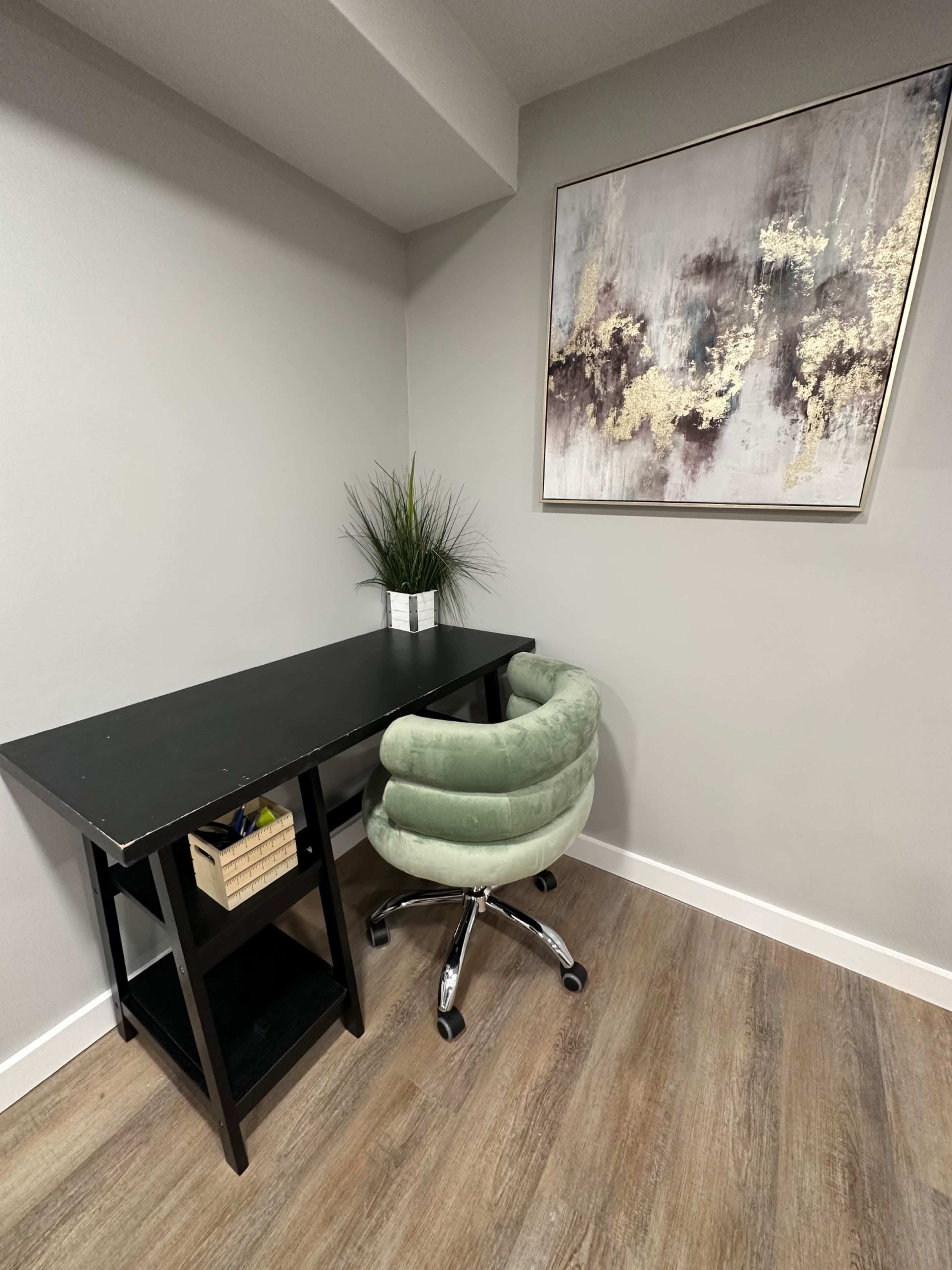 A minimalist workspace features a black desk with a green swivel chair and a potted plant, against a wall adorned with abstract art.