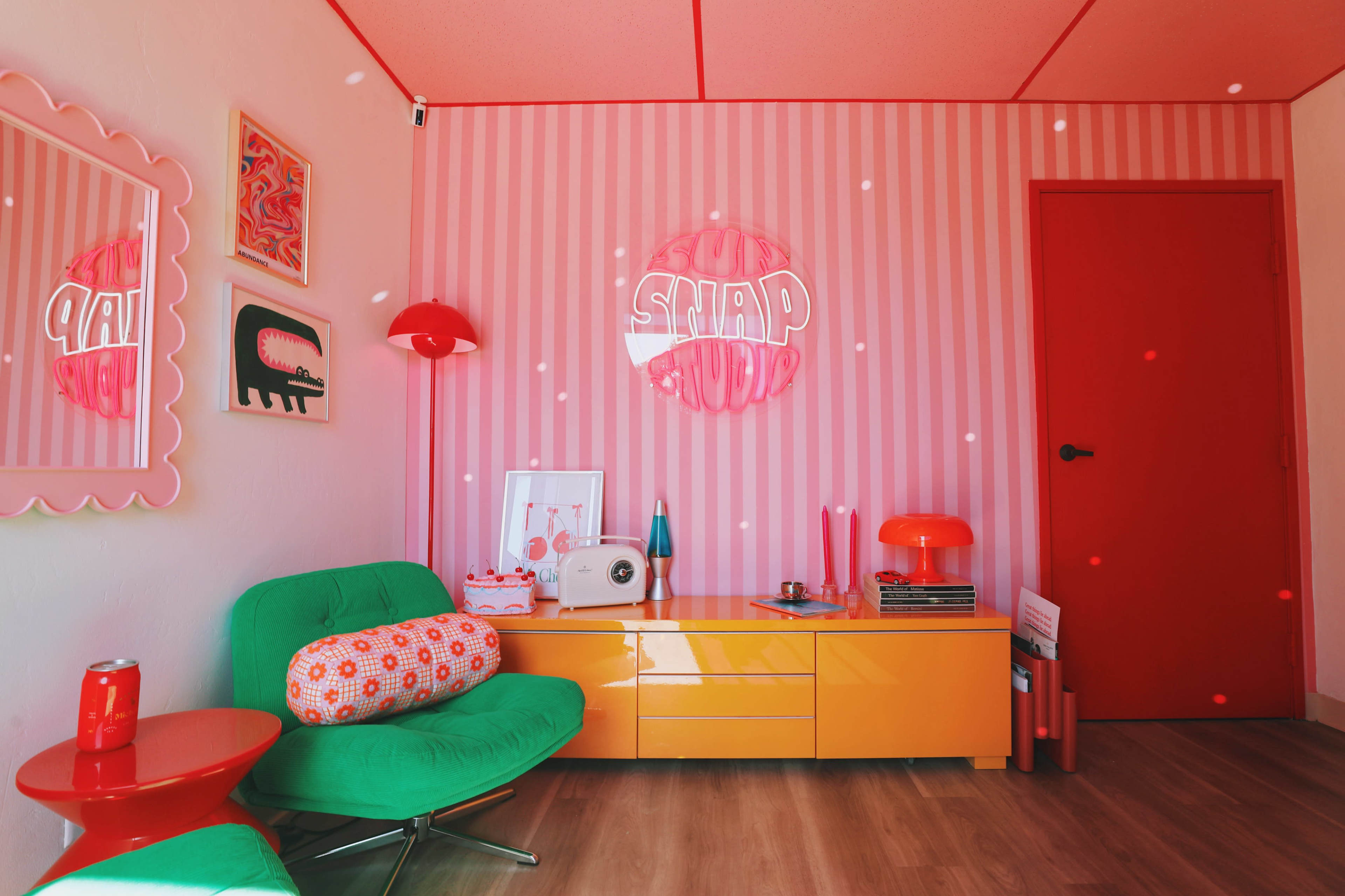 The room features colorful furniture and decor, with a green sofa, orange tables, and a neon sign, all against a backdrop of pink striped walls.
