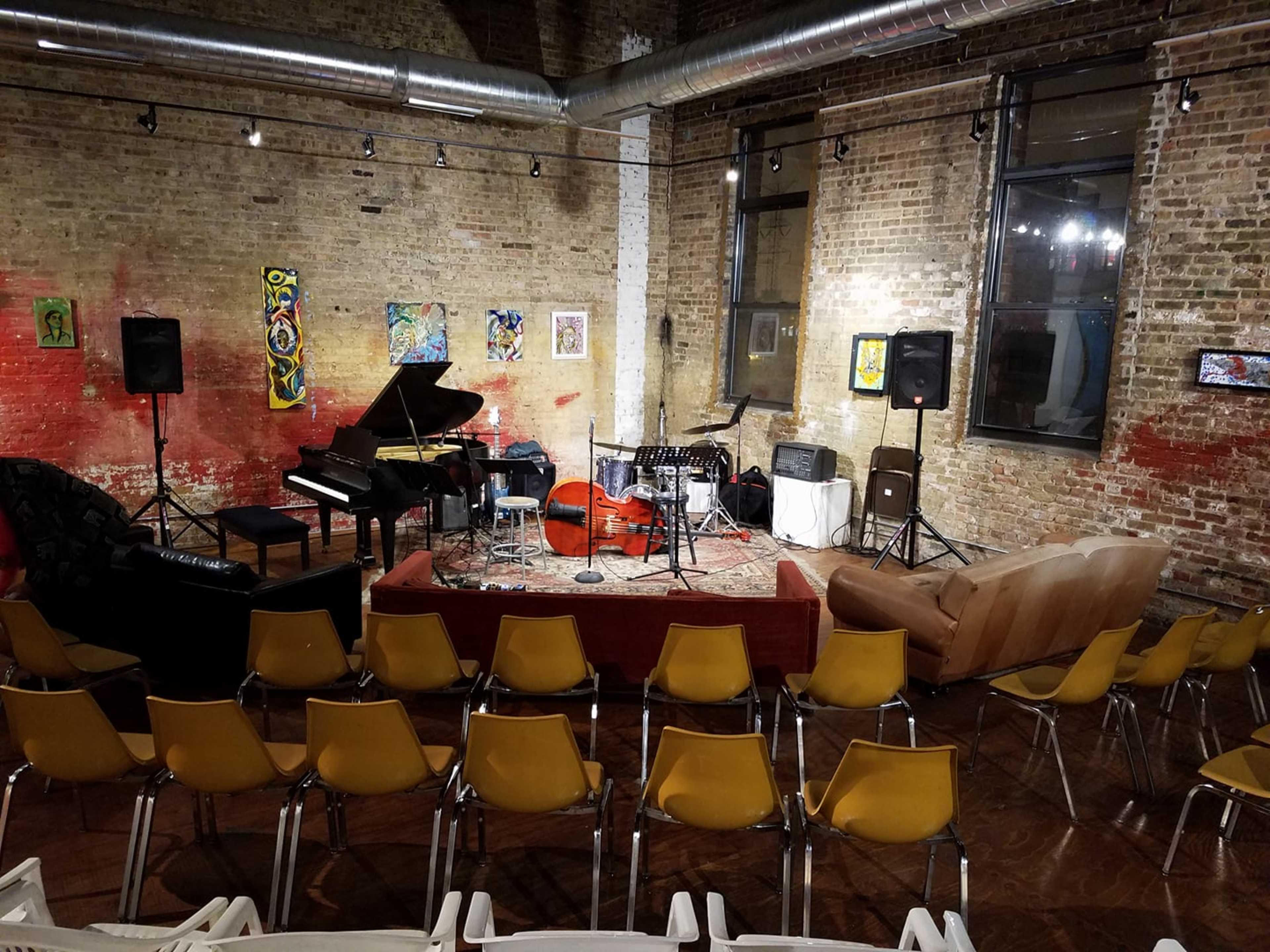 A small performance space features a piano, drum set, and string bass, arranged on a rug in front of chairs and a couch.