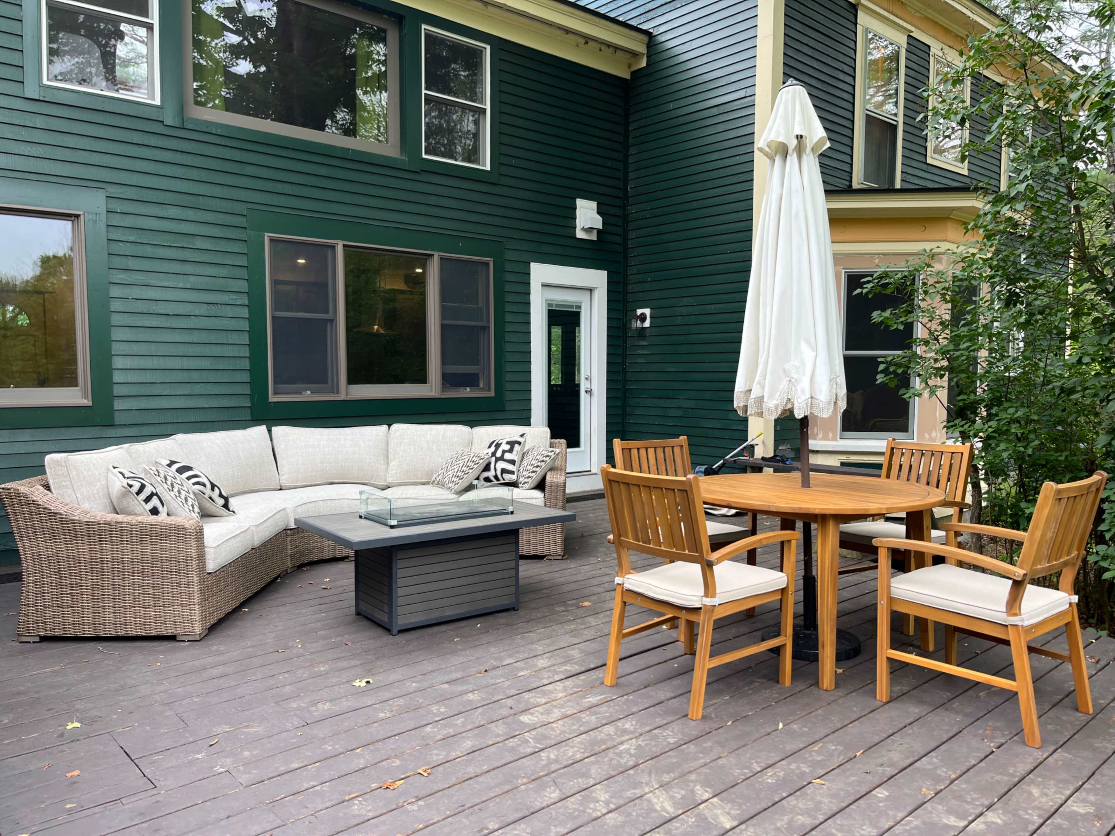 A cozy outdoor patio features a sectional sofa, a circular dining table with four chairs, and a large umbrella, all set against a green wooden house.