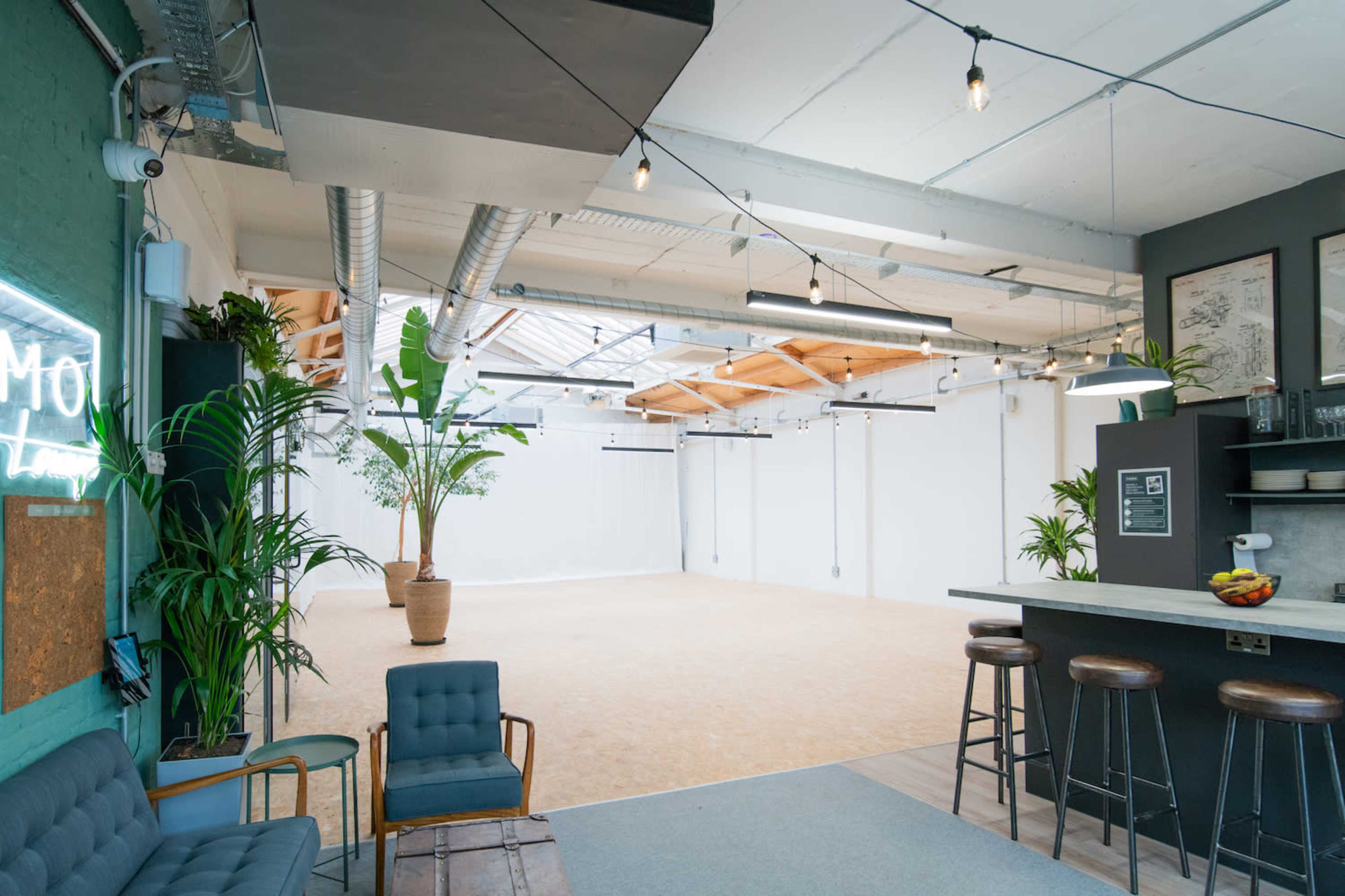 The image shows a spacious, well-lit interior space featuring greenery, wooden beams, and a minimalist setup with furniture along one side.