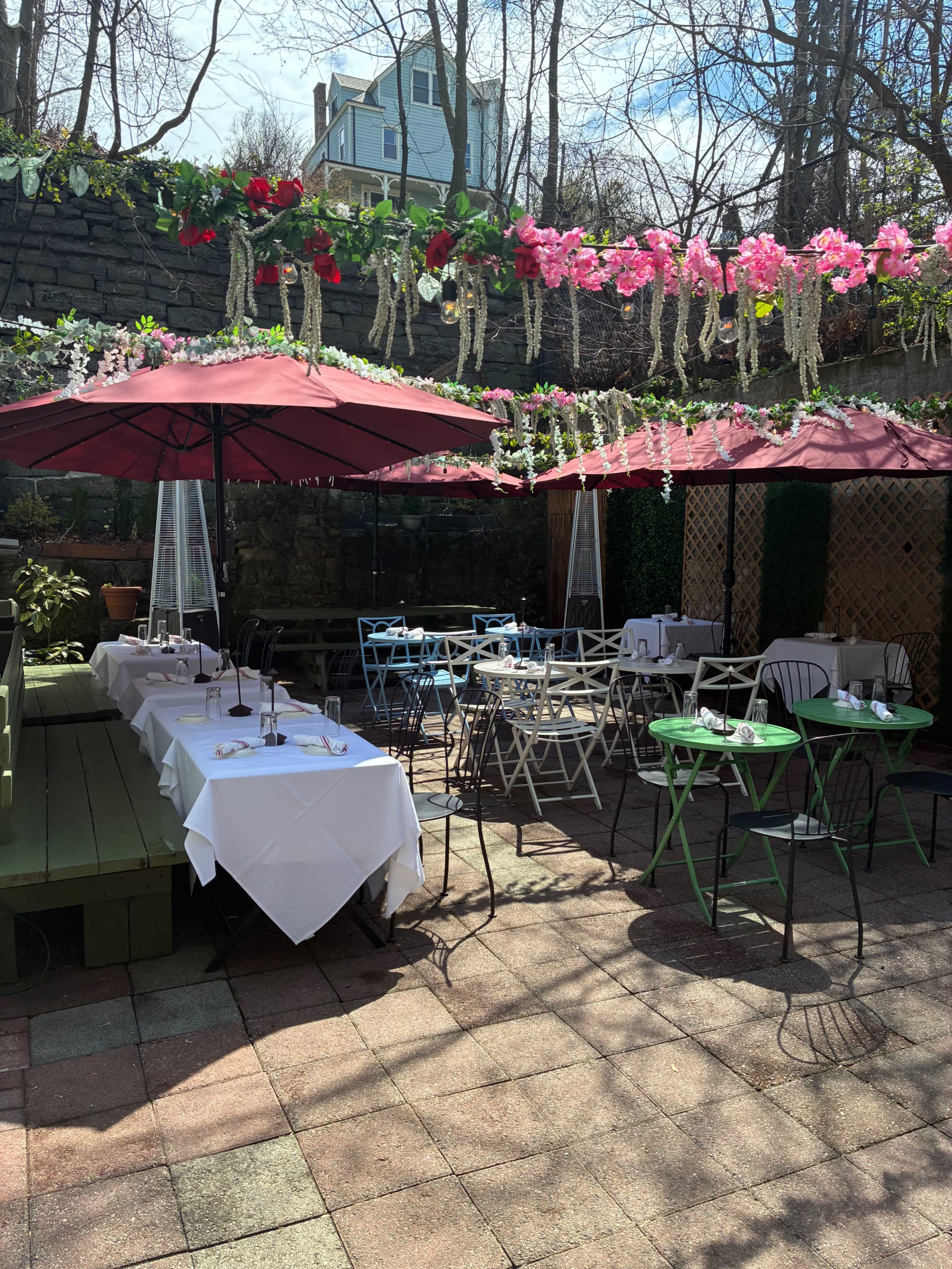 The image shows an outdoor dining area with tables and umbrellas, adorned with floral decorations and surrounded by greenery.
