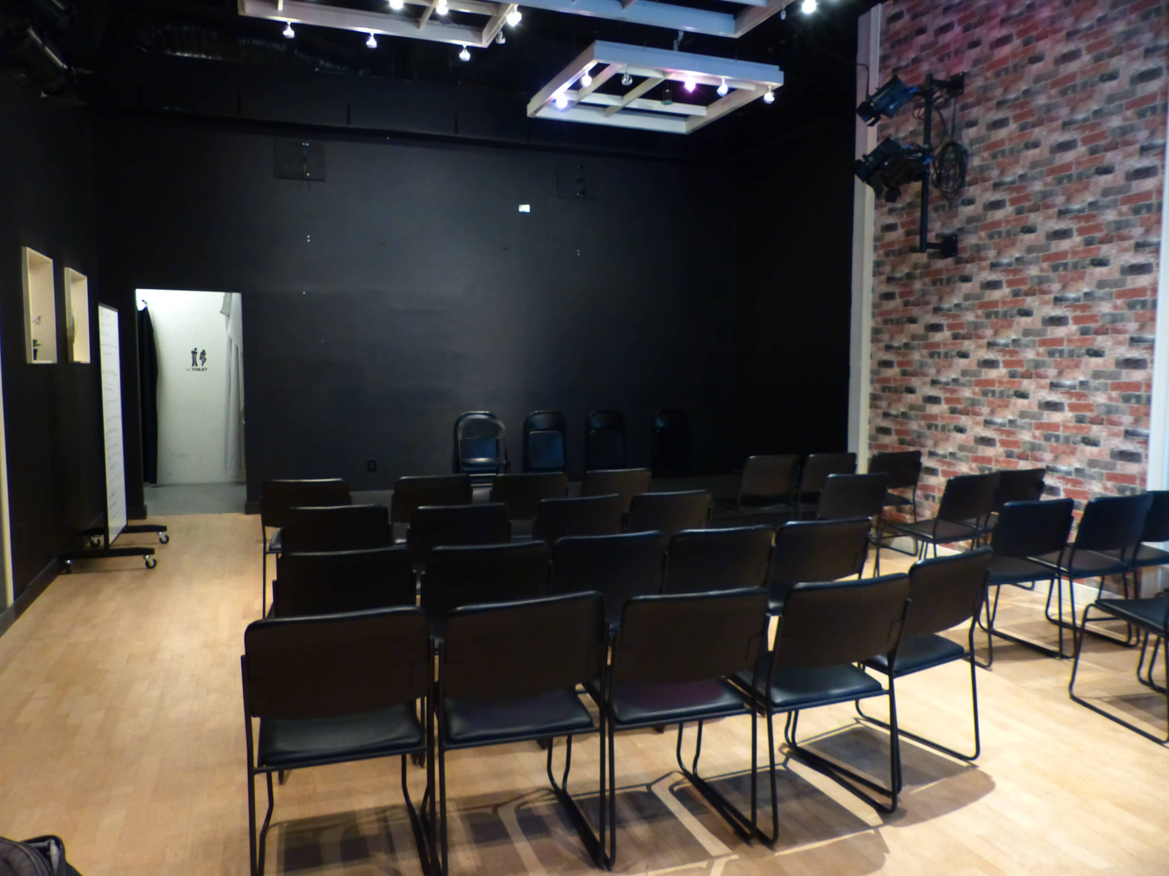 A small theater or meeting room with rows of black chairs facing a blank wall, and a doorway leading to a restroom area.