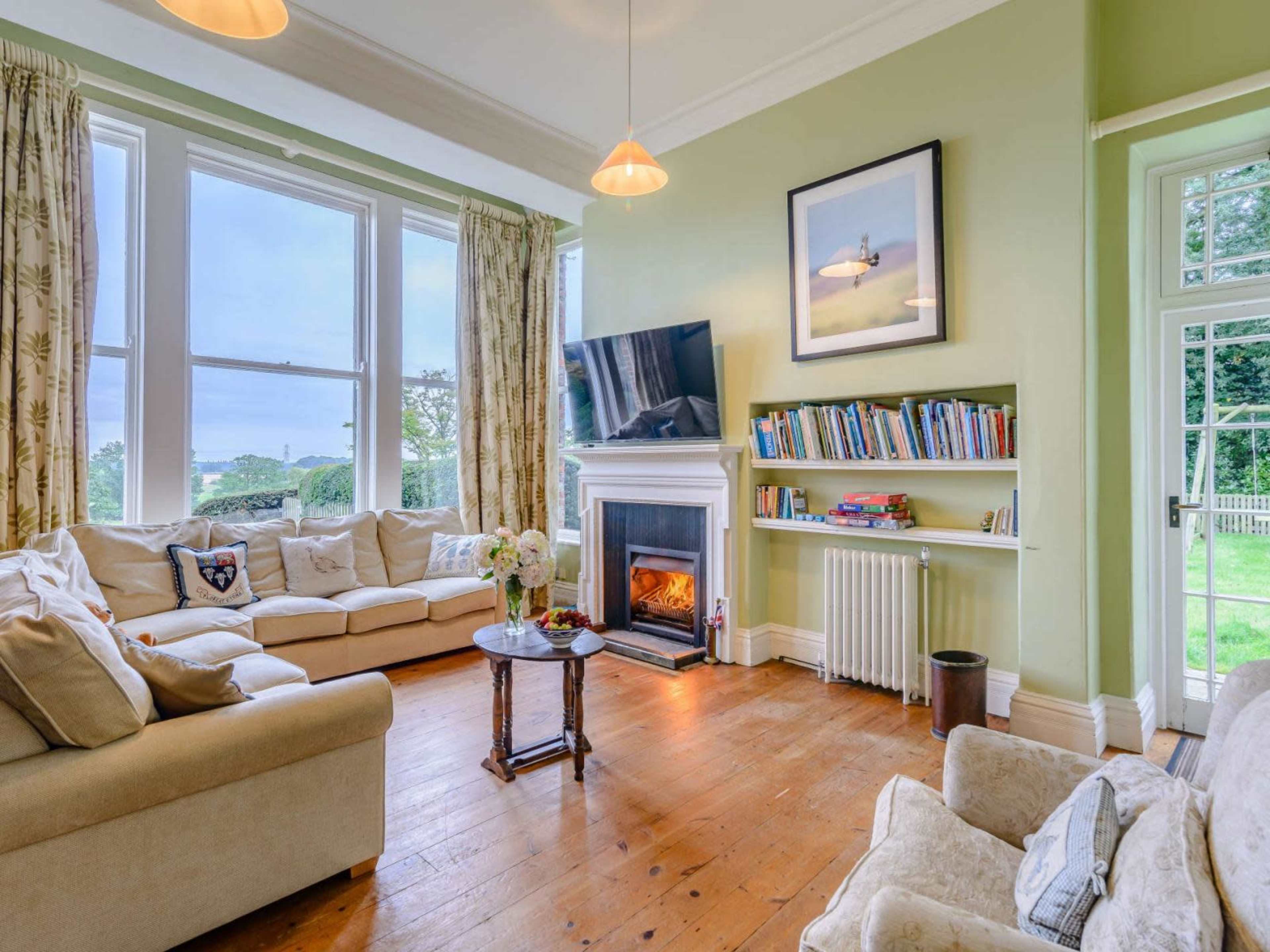 A cozy living room with a large window, a fireplace, a comfortable sofa, and a TV mounted on the wall above a shelf filled with books.