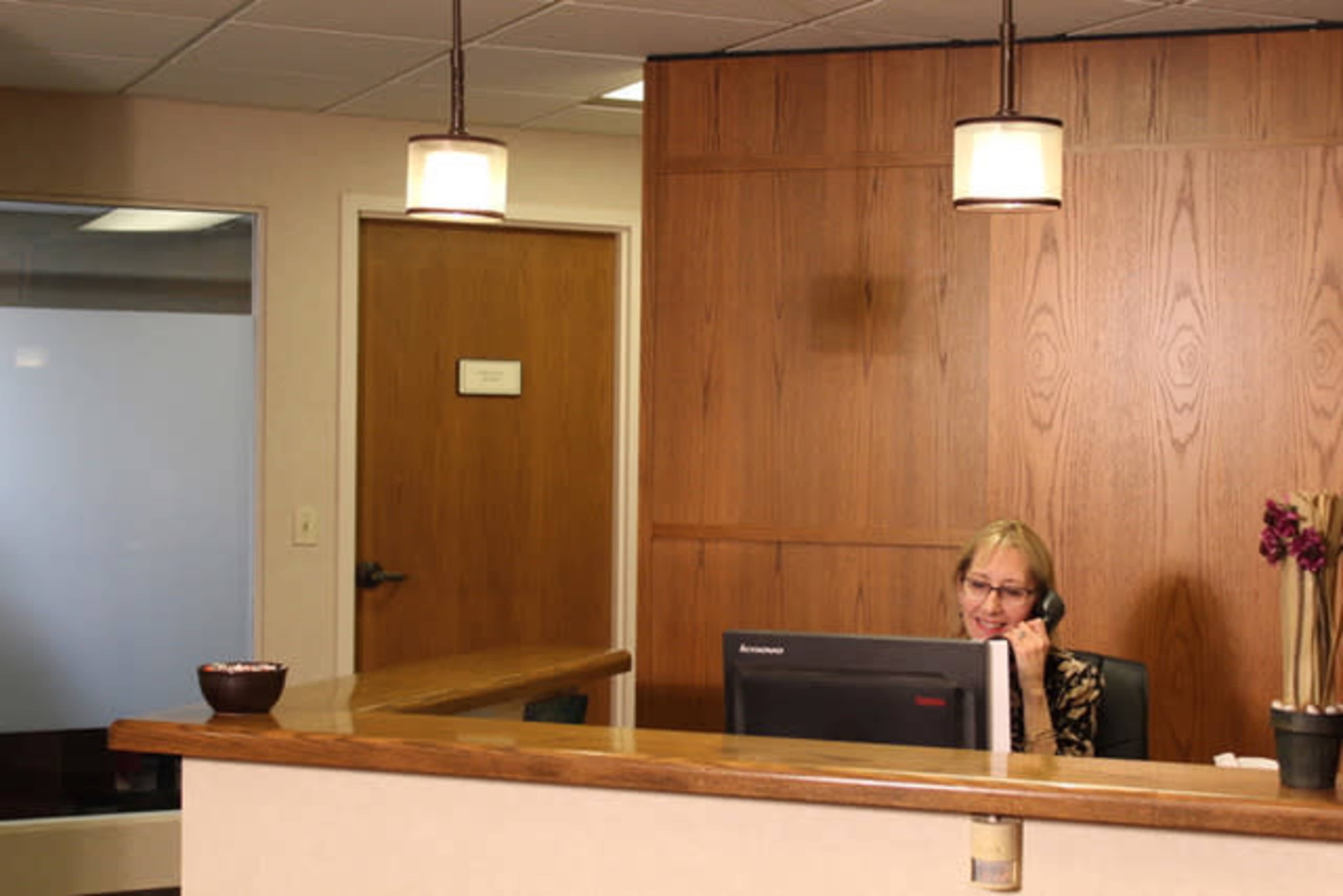 A receptionist sits at a desk, speaking on the phone in an office with wooden paneling and modern lighting.