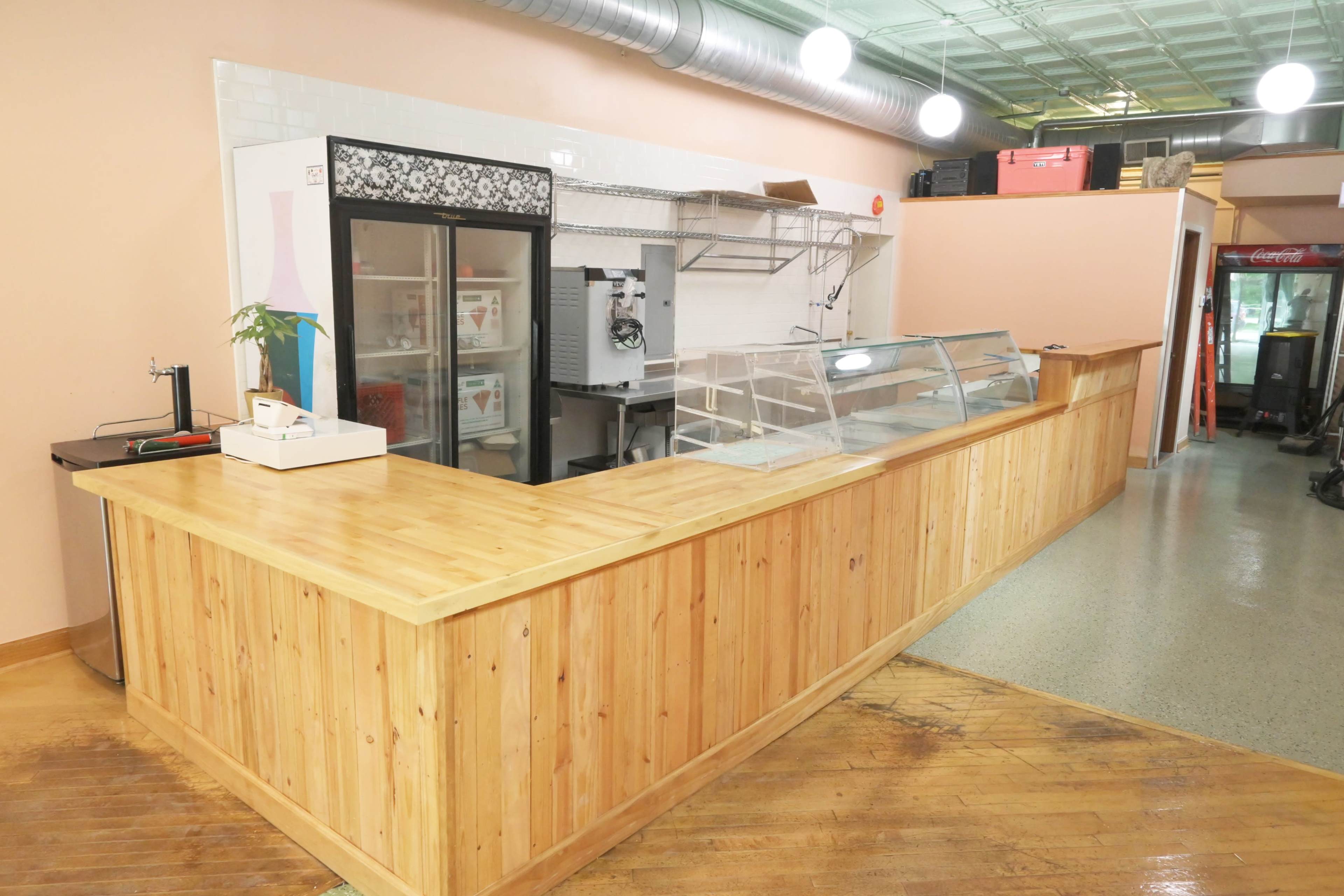 A wooden counter in an empty cafe with a display area, refrigeration units, and minimal decor.