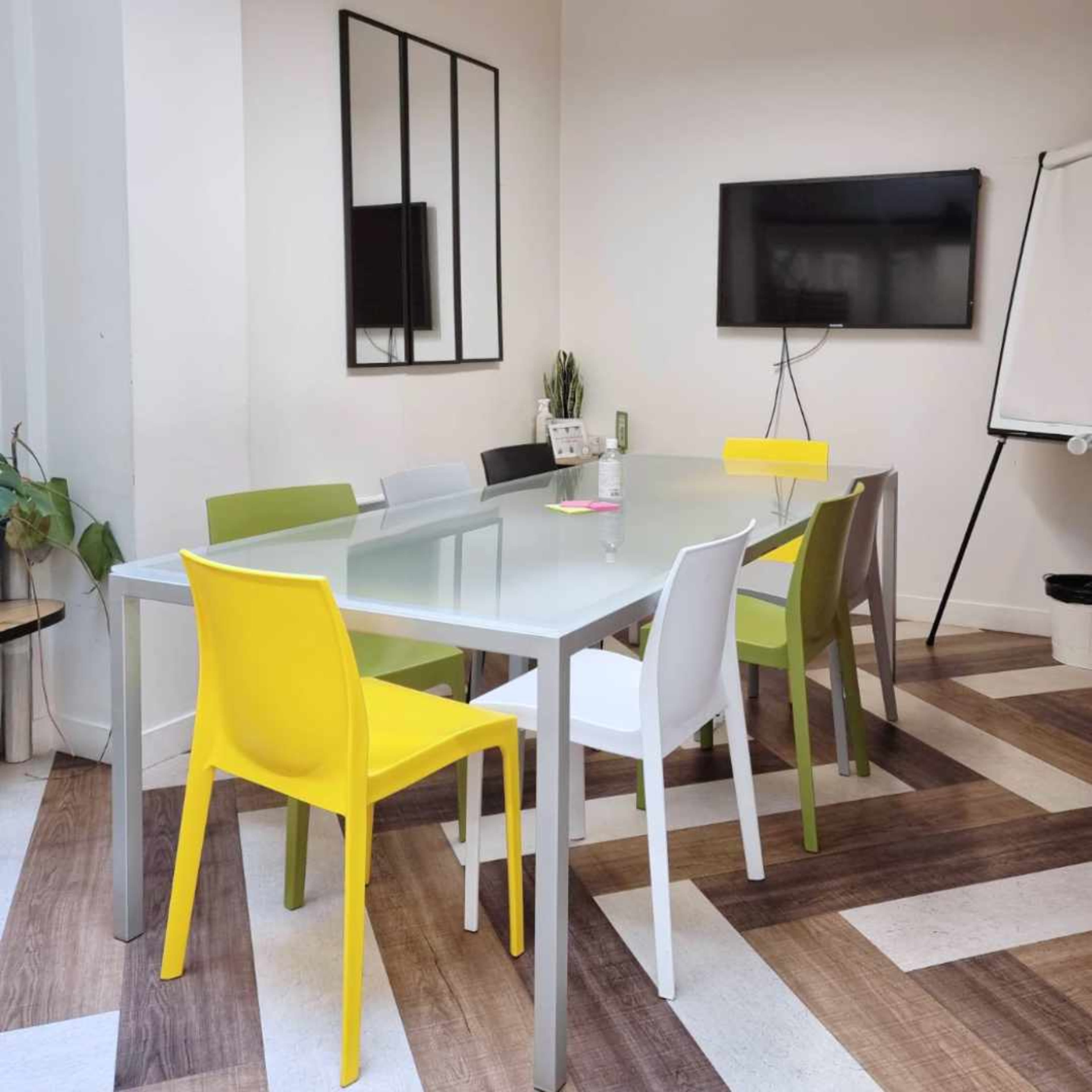 A meeting room features a glass table surrounded by six chairs in various colors, with a television and a whiteboard on the wall.