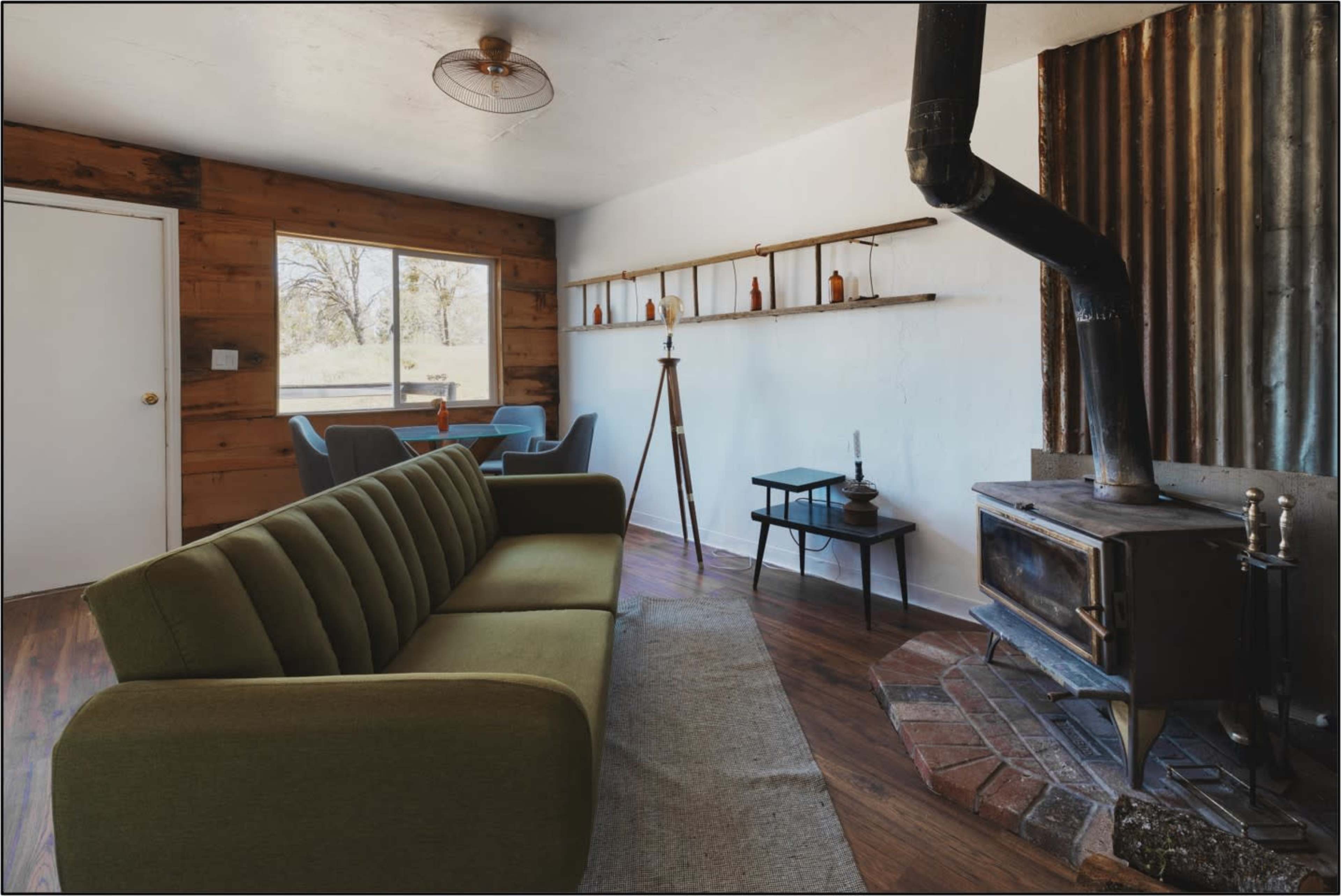 The room features a green sofa, a wooden dining area, and a metal stove against a rustic backdrop.