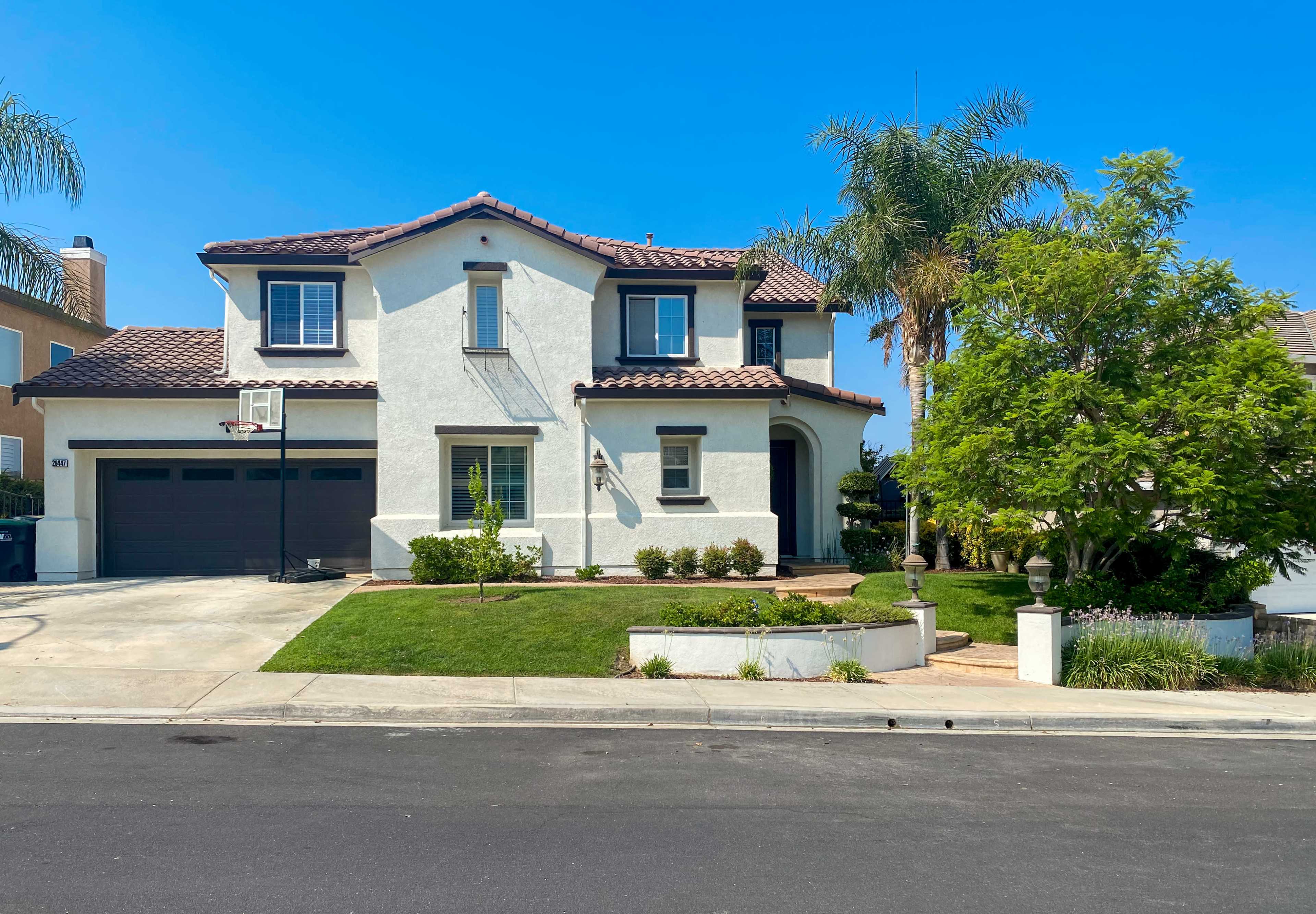 A two-story white house with a tile roof and a front yard featuring green grass and shrubs is located on a residential street.