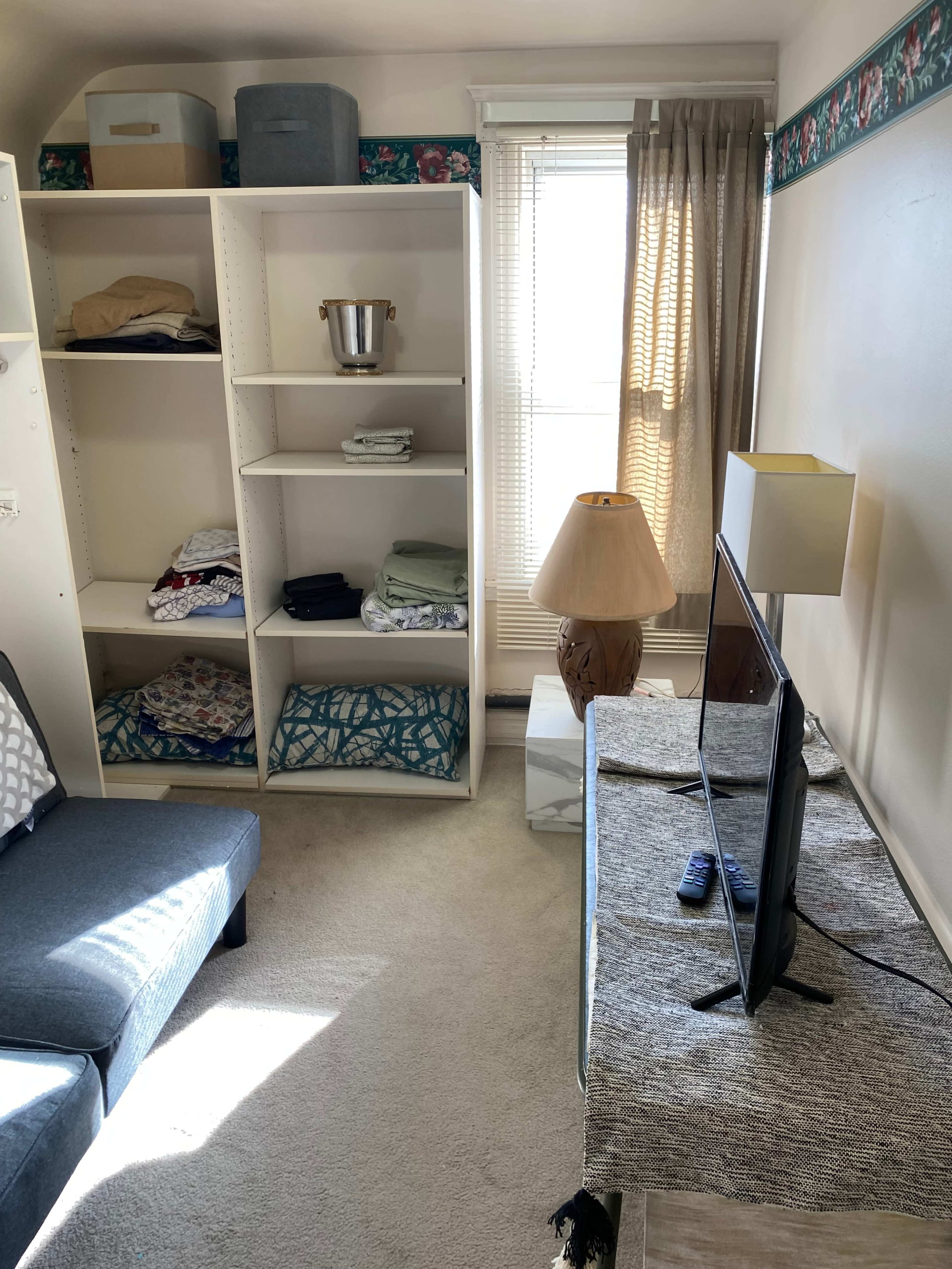 The image shows a small, well-organized room featuring a sofa, a television on a dresser, shelves with neatly folded clothing, and a lamp beside a window with curtains.