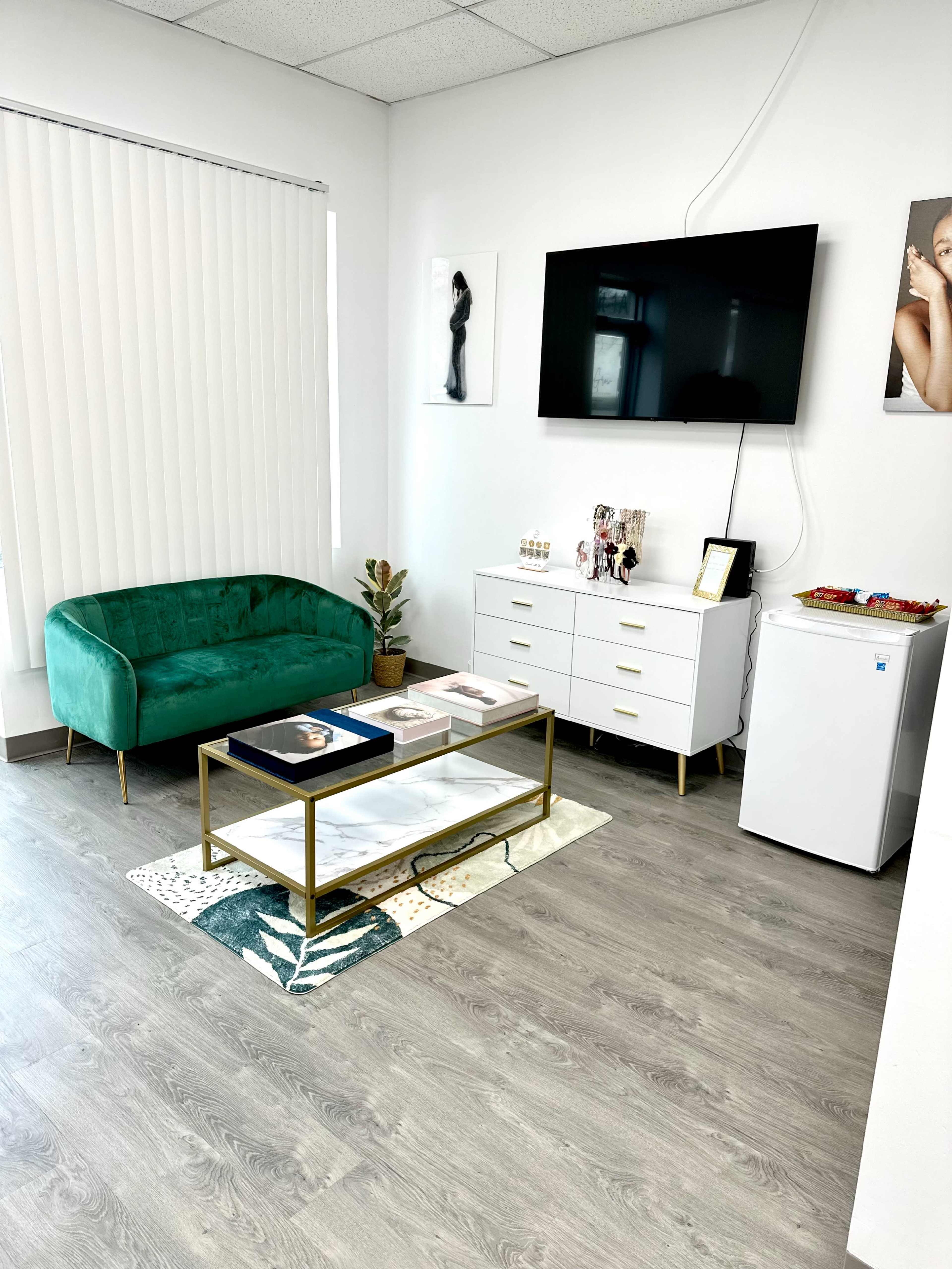 A modern waiting area with a green velvet couch, a wall-mounted television, a white dresser, and a small refrigerator, all arranged on a light wood floor.