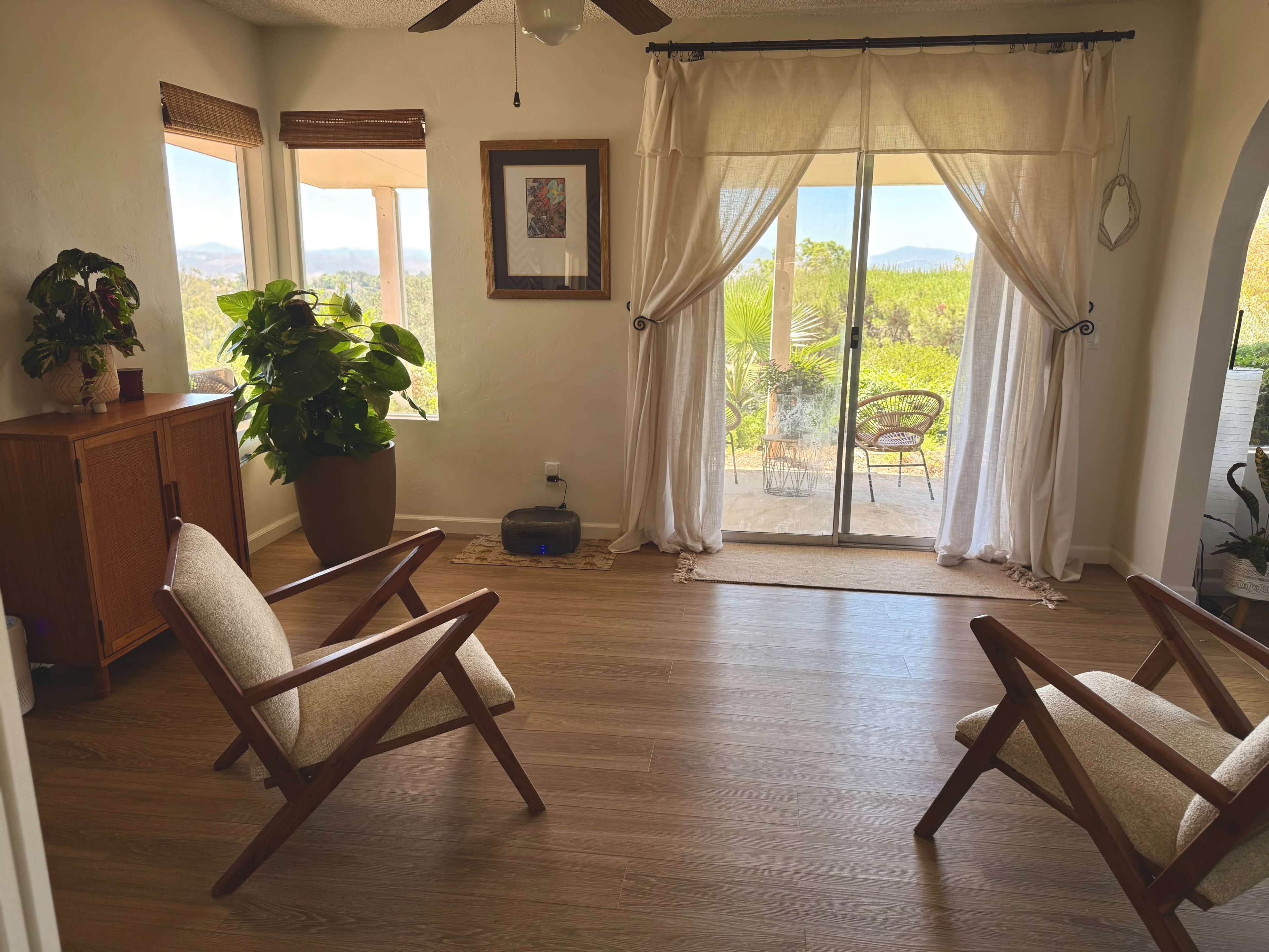 A bright room features two wooden chairs facing sliding glass doors that open to a view of greenery and hills.