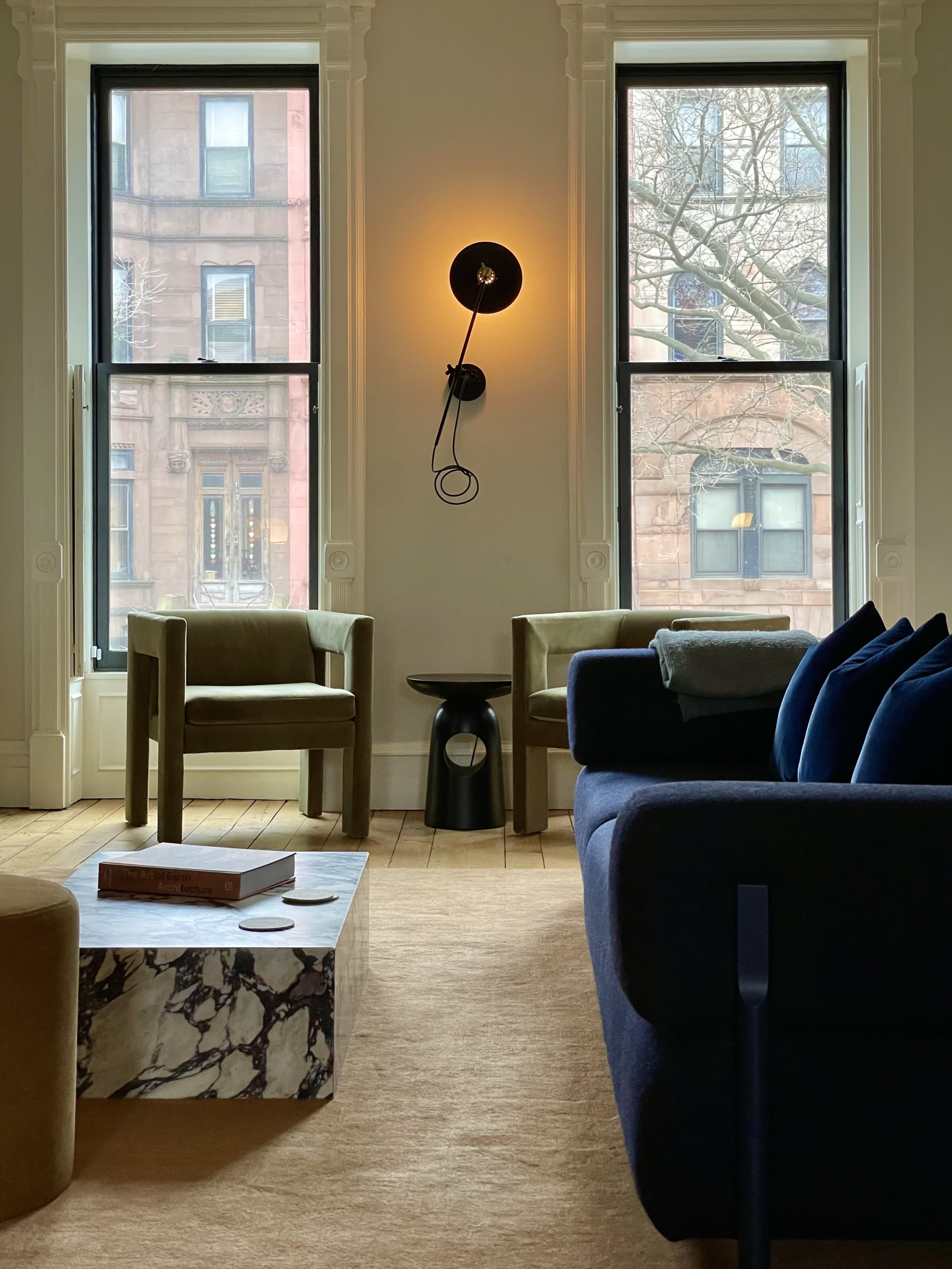 A modern living room with two green chairs, a marble coffee table, and a blue couch, all positioned near large windows with a view of a red brick building outside.