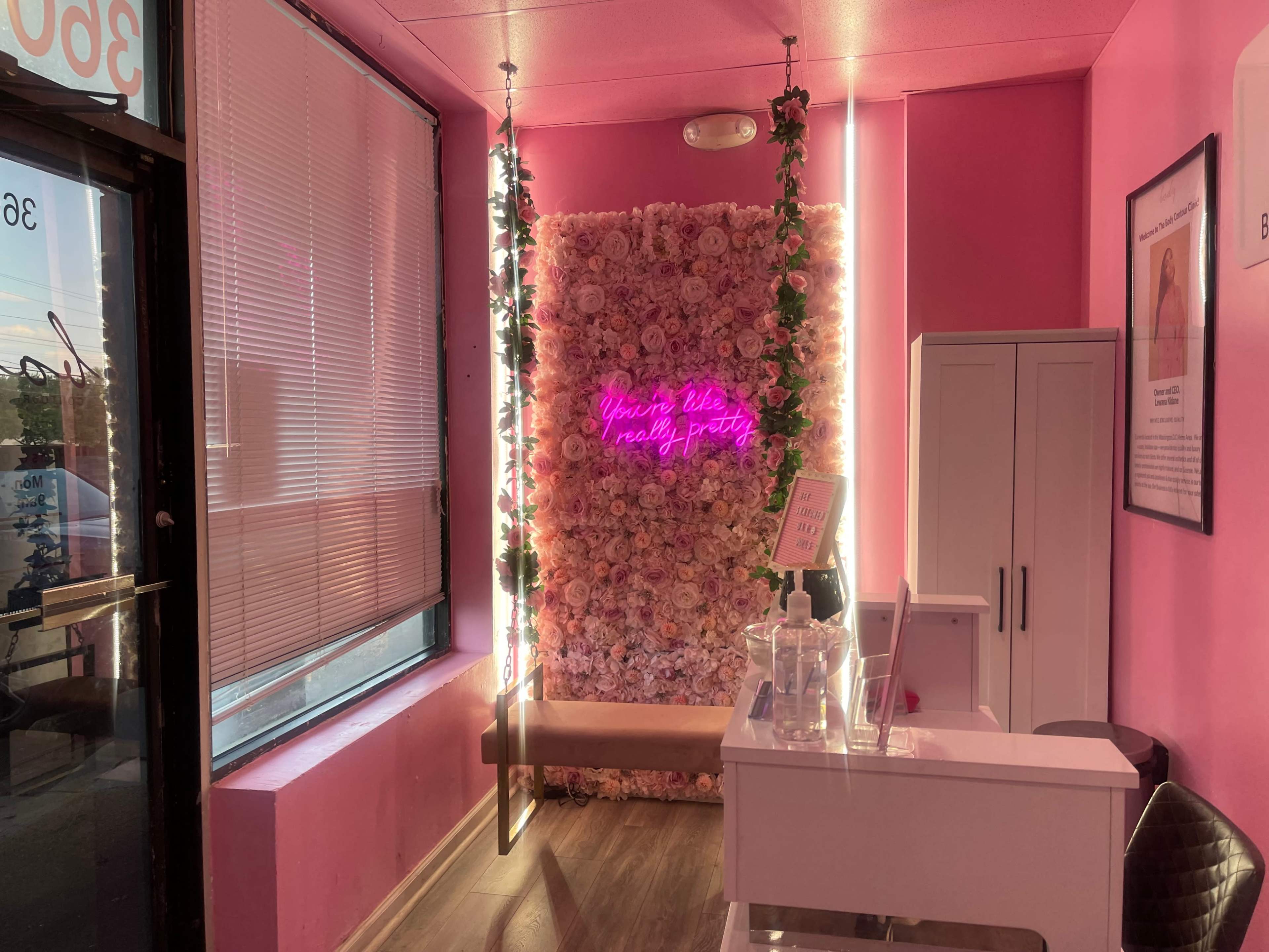 A pink-themed interior with a wall covered in artificial flowers, featuring a neon sign that reads "You're like really pretty."
