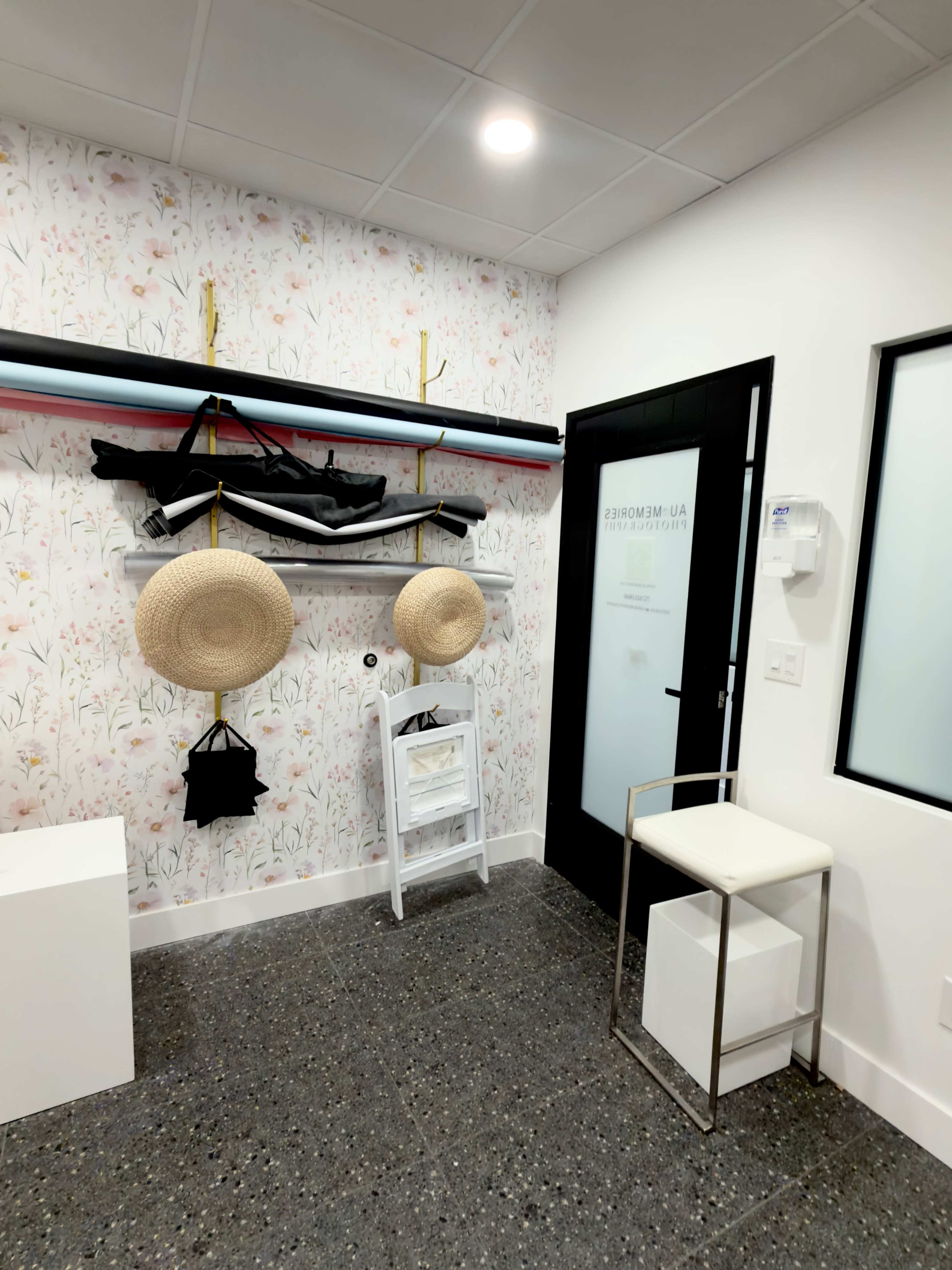 The image shows a modern entryway with floral wallpaper, a black door, and a wall-mounted rack supporting items like surfboards and hats.